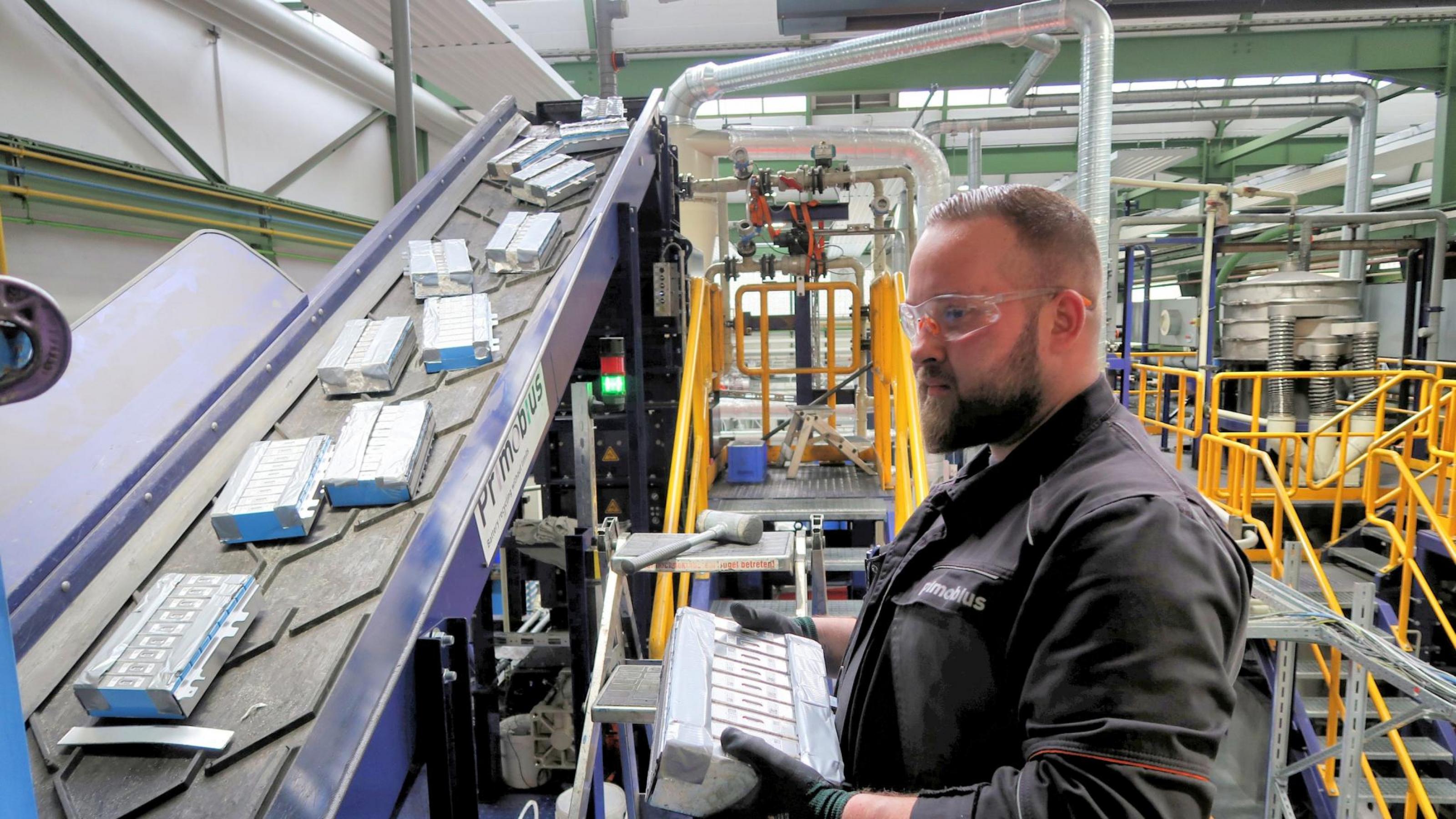 Ein Mitarbeiter mit Schutzbrille steht an einem Fließband mit Autobatterien in einer Werkshalle.