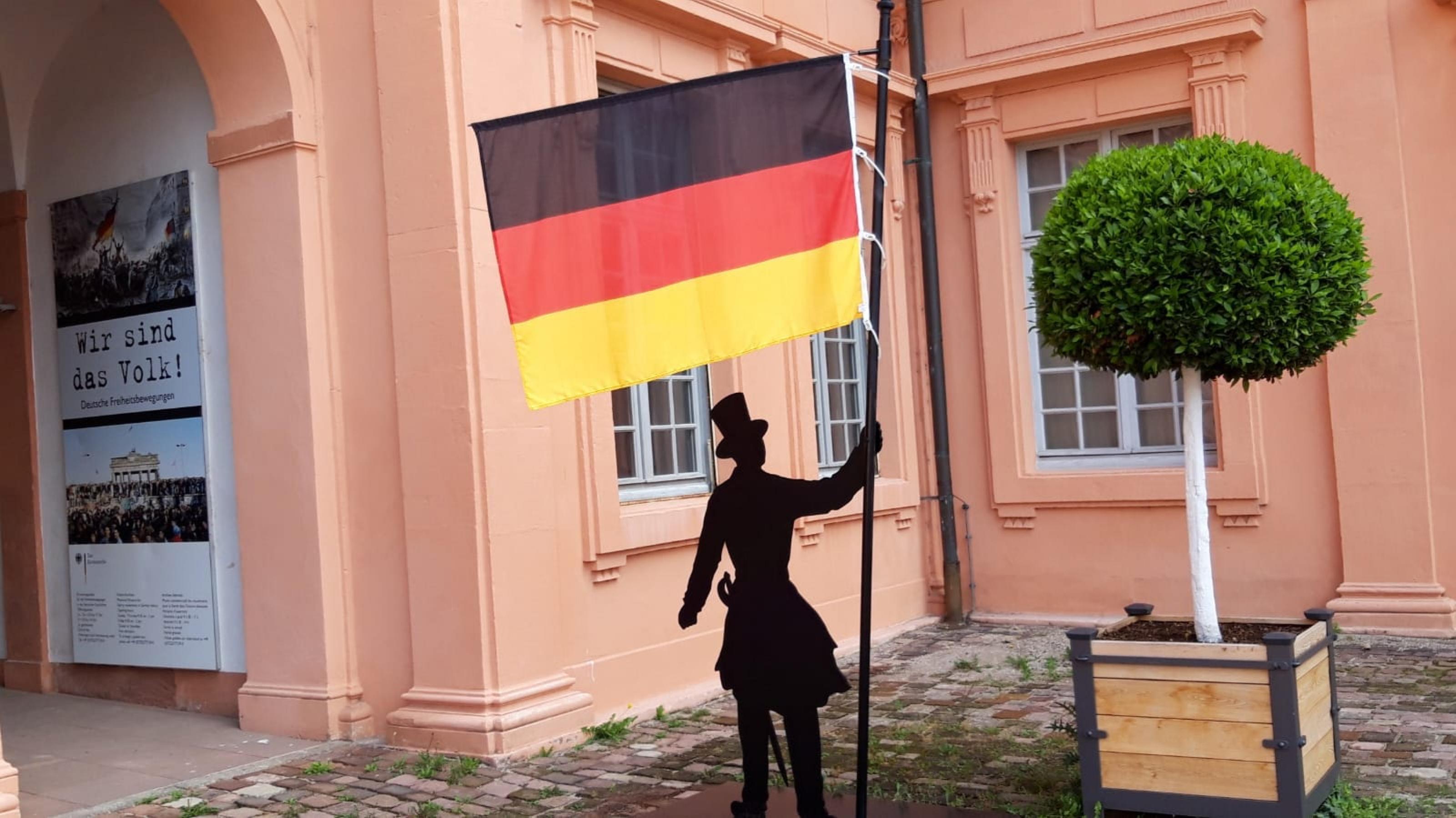 Ein Museums-Hinweis-Männchen mit Deutschlandflagge vor dem rostroten Sansteingebäude.