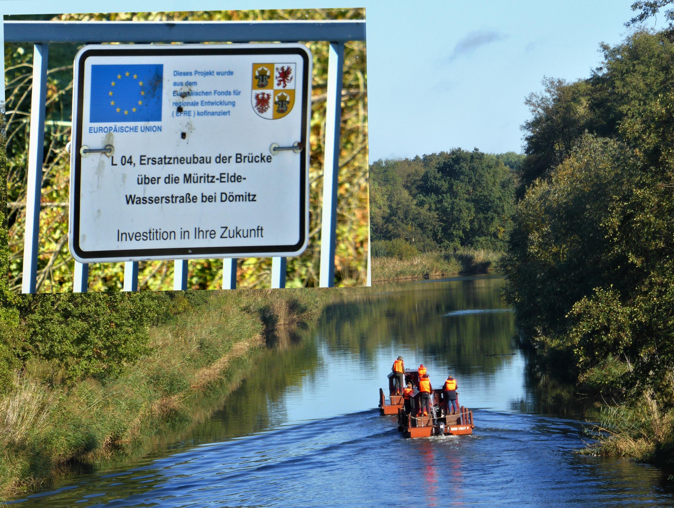 Unter blauem Himmel und auf blauem Wasser schippern zwei floßartige Wassergefährte mit jeweils zwei Personen Besatzung weg von Betrachter/-in. Zusätzlich trägt das Foto ein Insert, ein Ausschnitt vom Brückengeländer, das die EU-Förderung des „Brücken-Ersatzneubaus“ belegt.