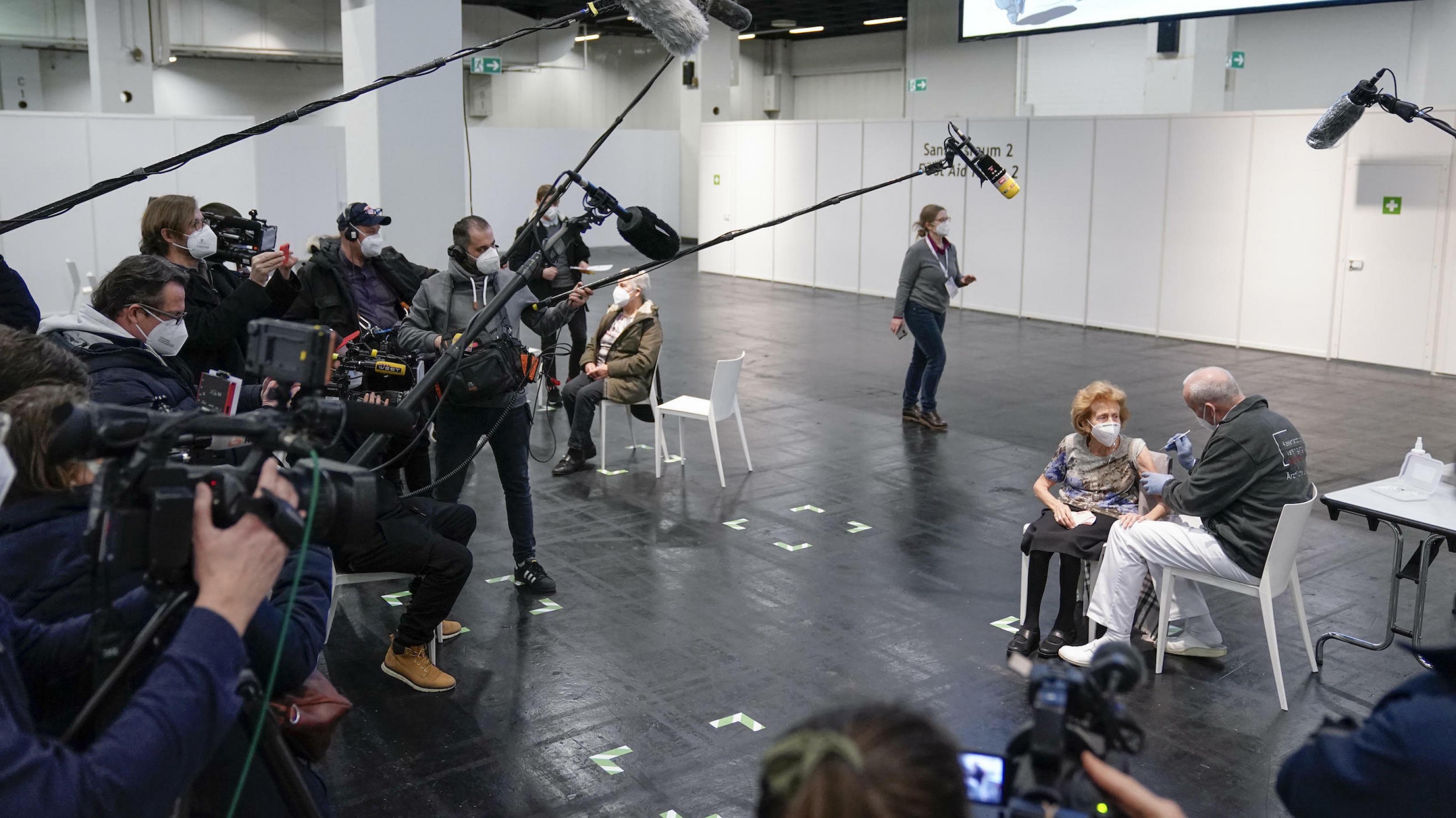 In einer großen Halle: Menschen mit Kameras und Mikrofonen umringen eine ältere Frau auf einem Stuhl, die sich von einem Mann eine Spritze in den rechten Oberarm verabreichen lässt.