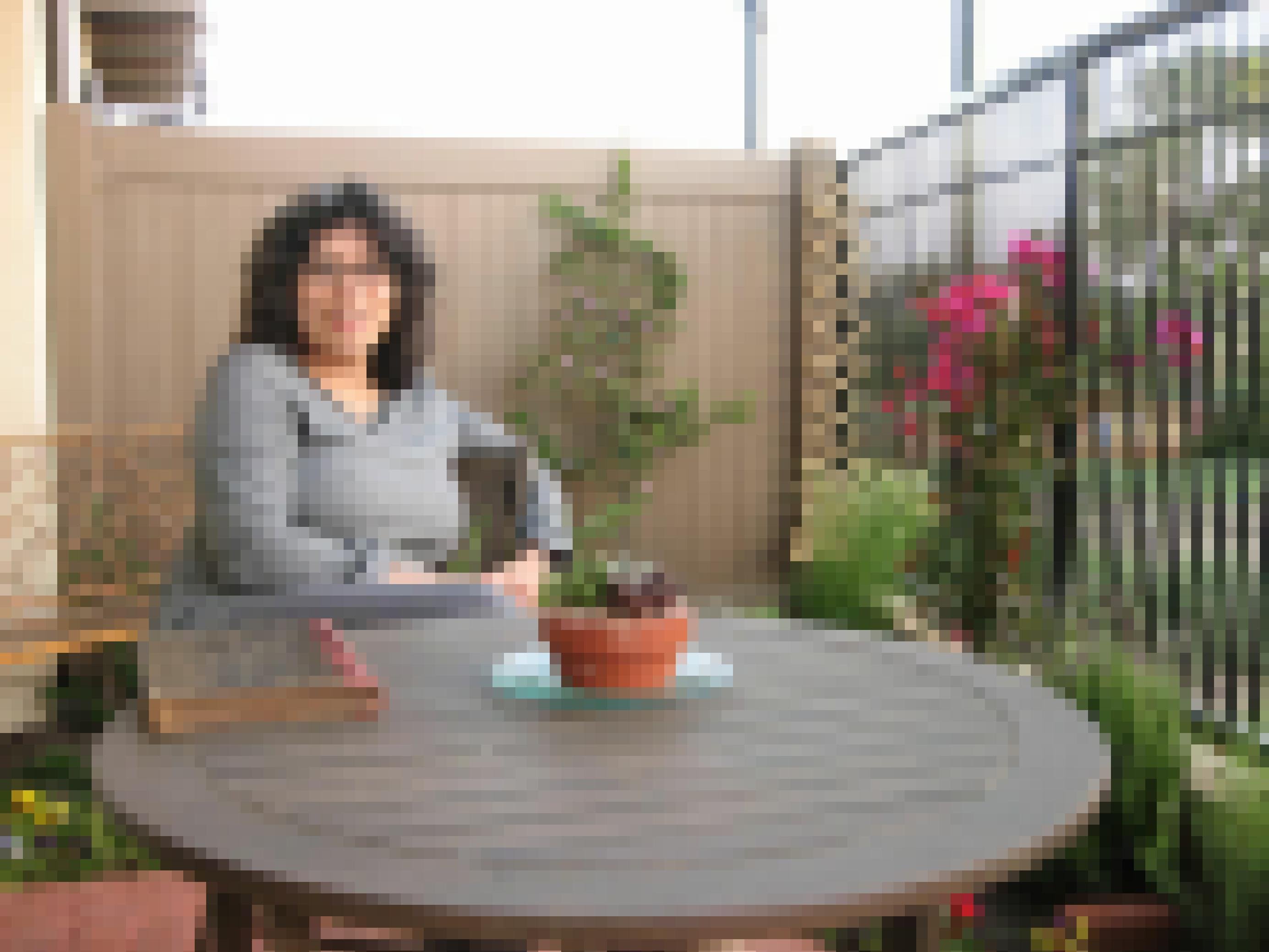 Eine Terrasse mit einem runden Holztisch, dahinter eine Holzwand, an der sich eine Pflanze hochrankt. Am Tisch sitzt eine rund 40-jährige Frau mit dunklen Locken, grosser Brille und grauem Pullover. Vor ihr auf dem  Tisch ein alter Foliant.