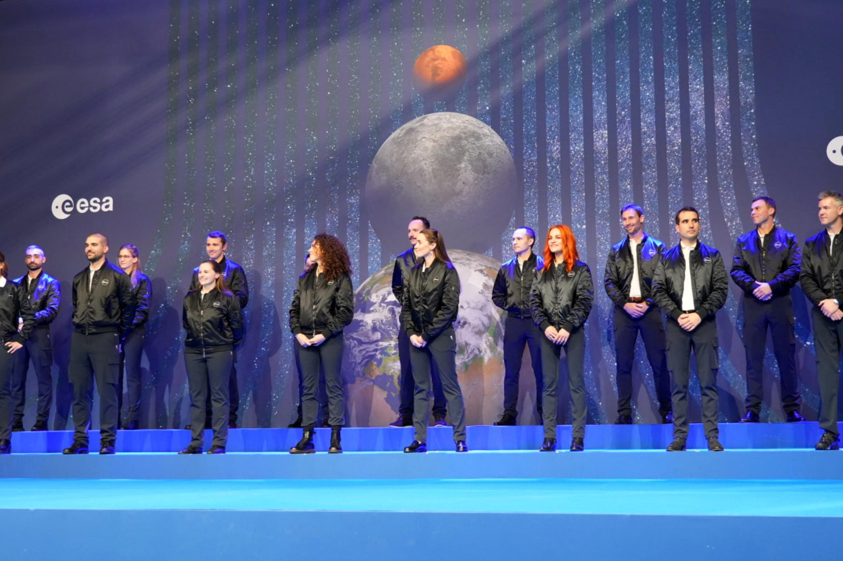 Eine Bühne mit Menschen in schwarzen Jacken. Hinter ihnen eine Leinwand mit einem Mond und einem roten Planeten und dem ESA-Logo.