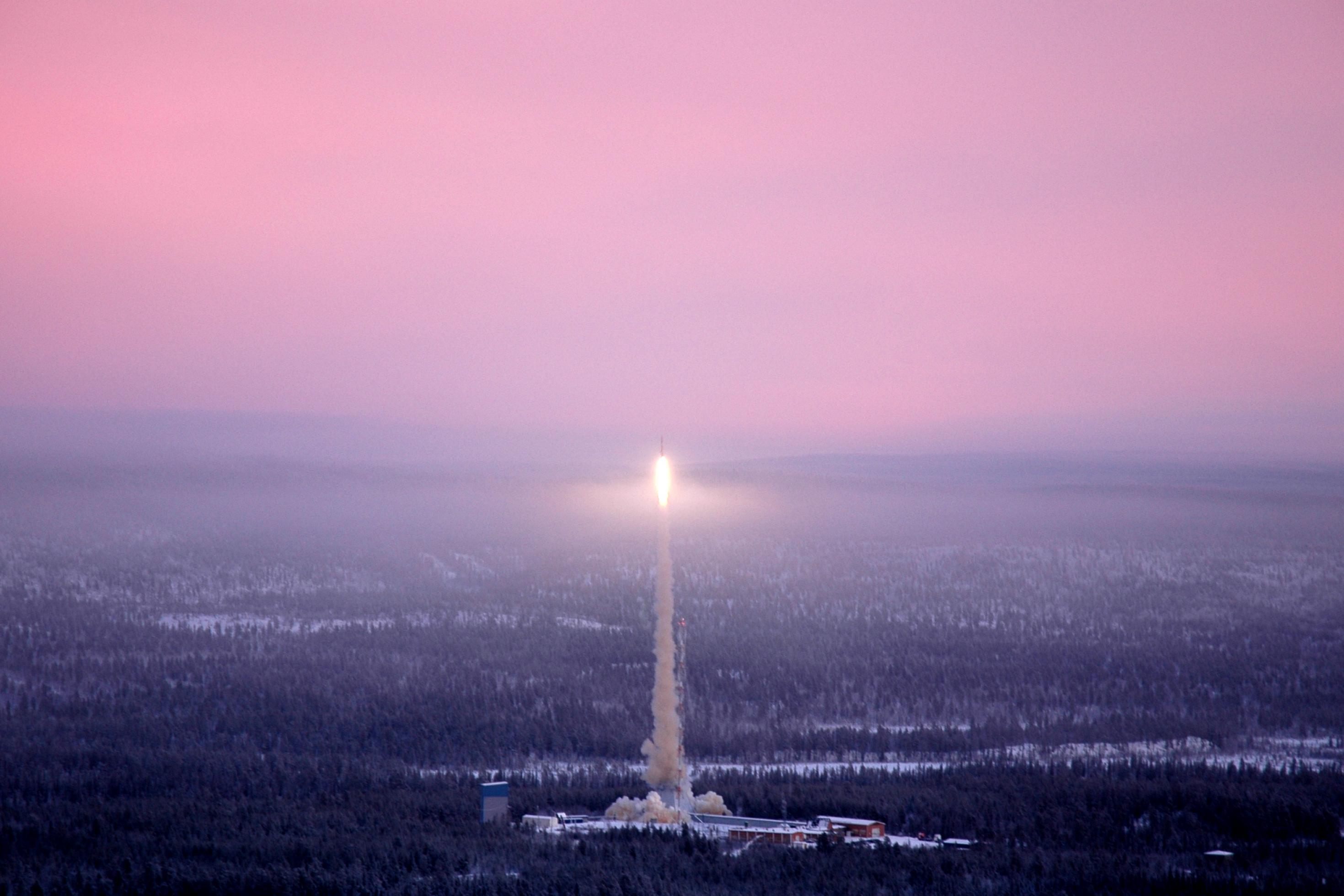 Luftbildaufnahme einer verschneiten, dunstigen Landschaft mit Wald, im Zentrum erstrahlt das Triebwerk einer Rakete im Flug.