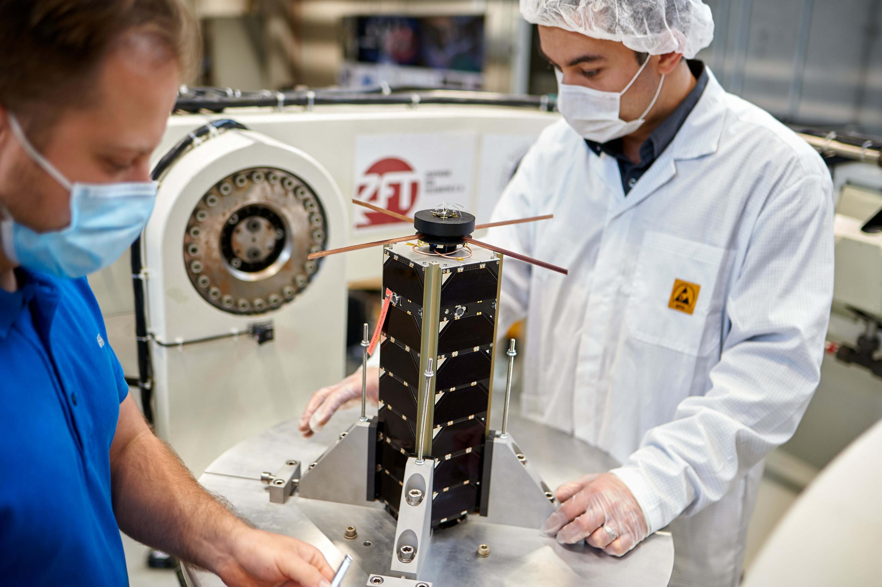Einer der NetSat-CubeSats steht im Reinraum: etwa so groß wie ein Schuhkarton mit Solarzellen auf den Außenflächen, daneben unscharf zwei Herren im Ganzkörperschutz.