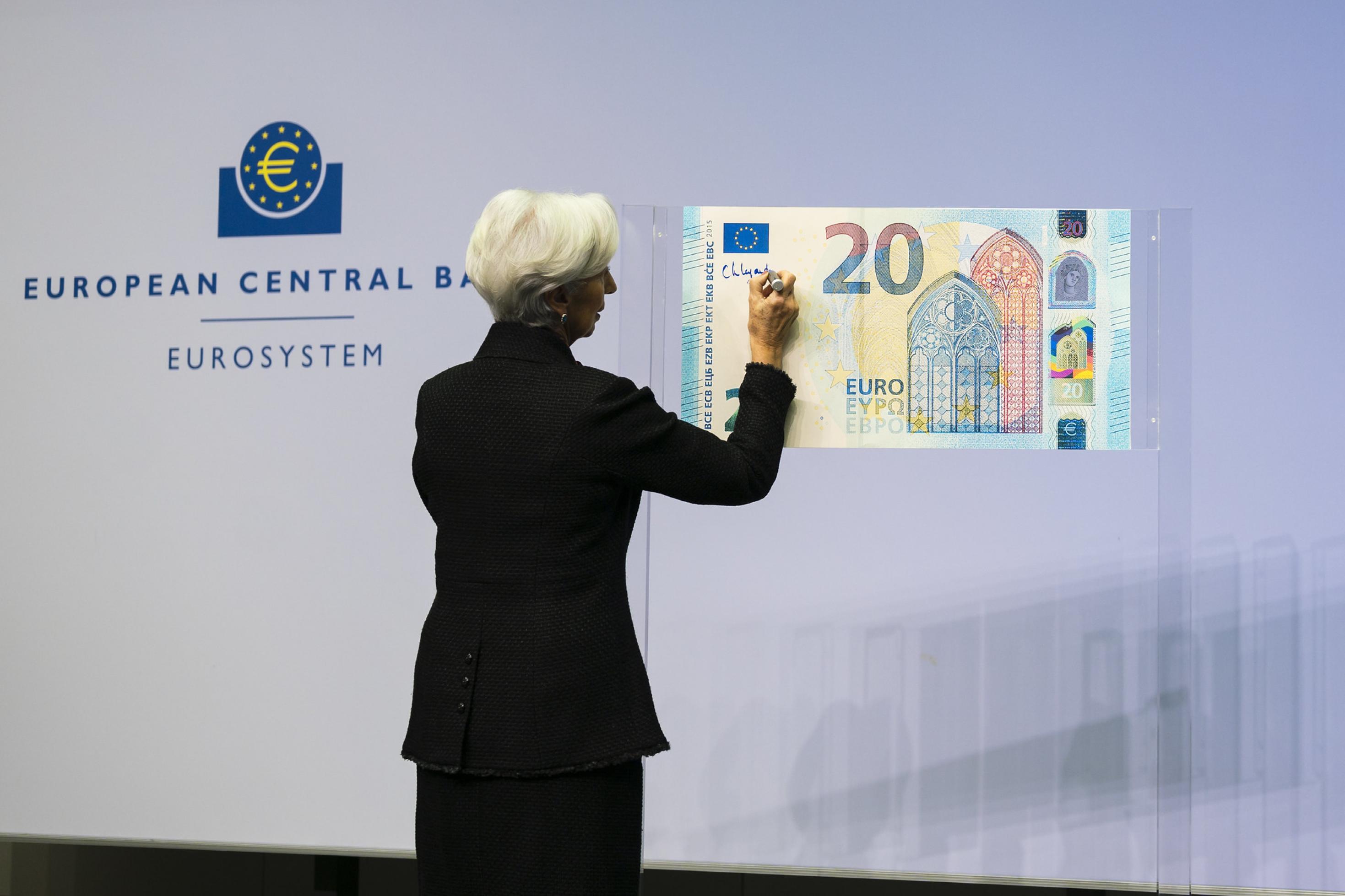 Lagarde, eine Frau mit weißem Haar, unterschreibt mit dickem Stift eine große, an die Wand gehängte Banknote.