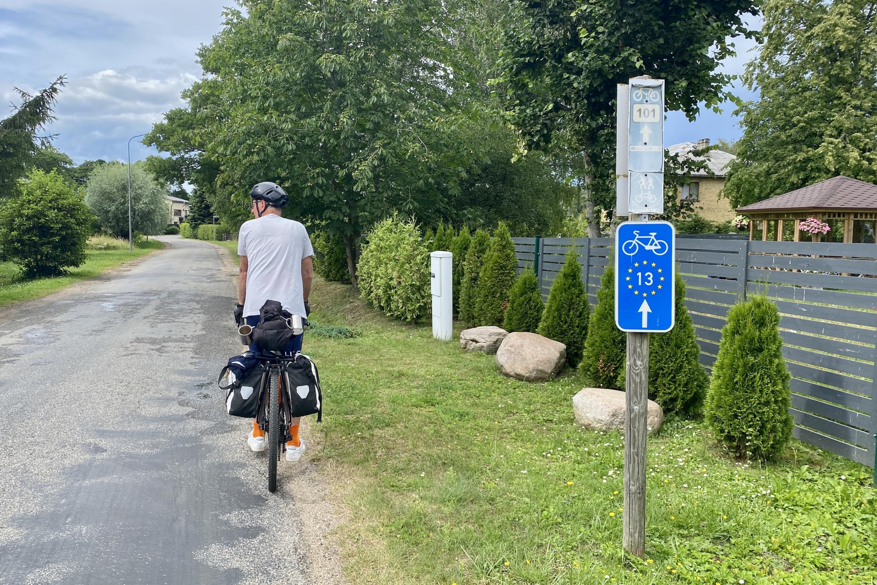Ein Radfahrer mit Fahrradtaschen steht am Fahrbahnrand. Ein Schild markiert den Weg als Eurovelo-Route 13.