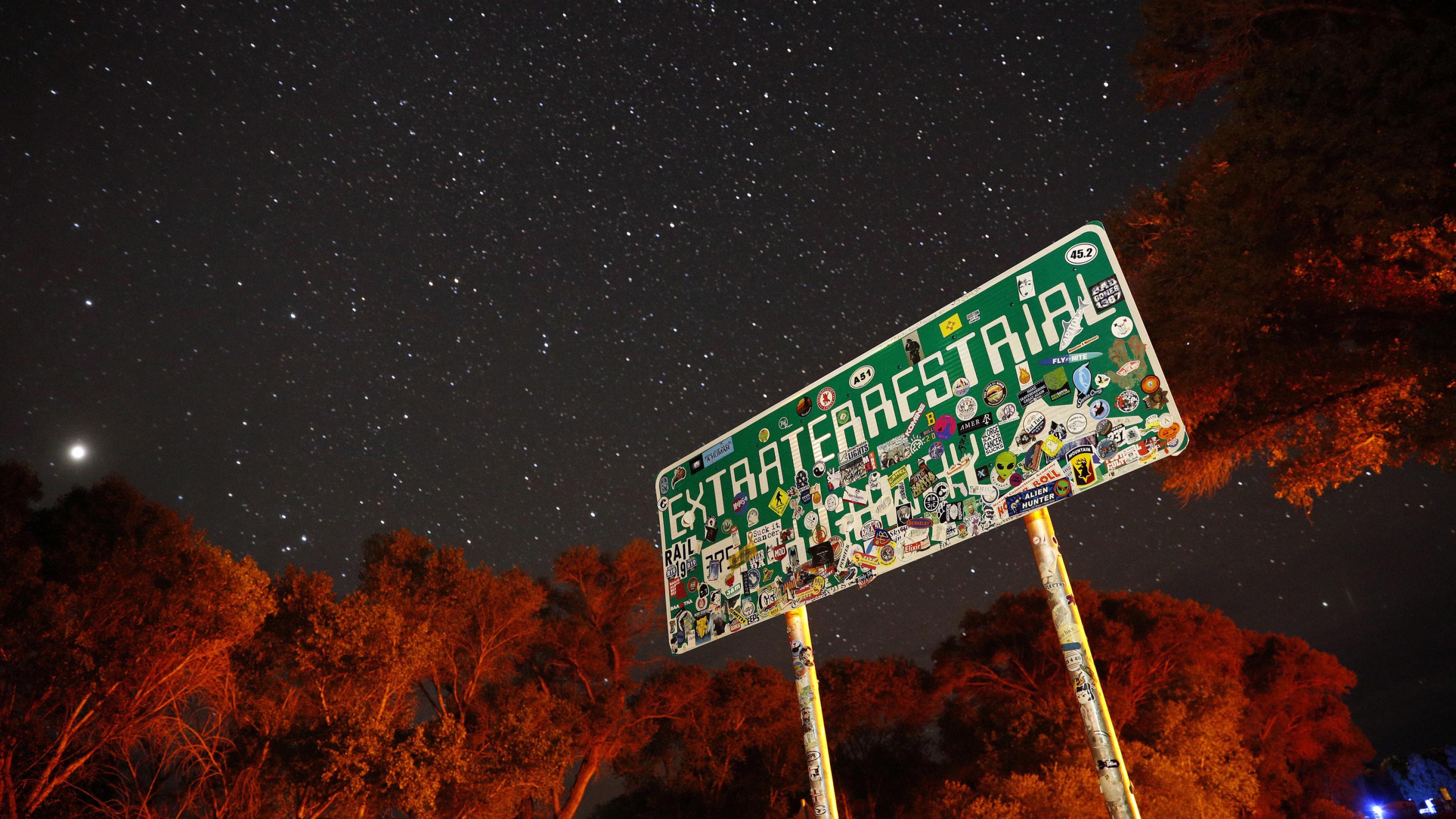 Ein mit Aufklebern überklebtes Schild mit der Aufschrift „Extraterrestrial Highway“ steht nachts unter einem Sternenhimmel.