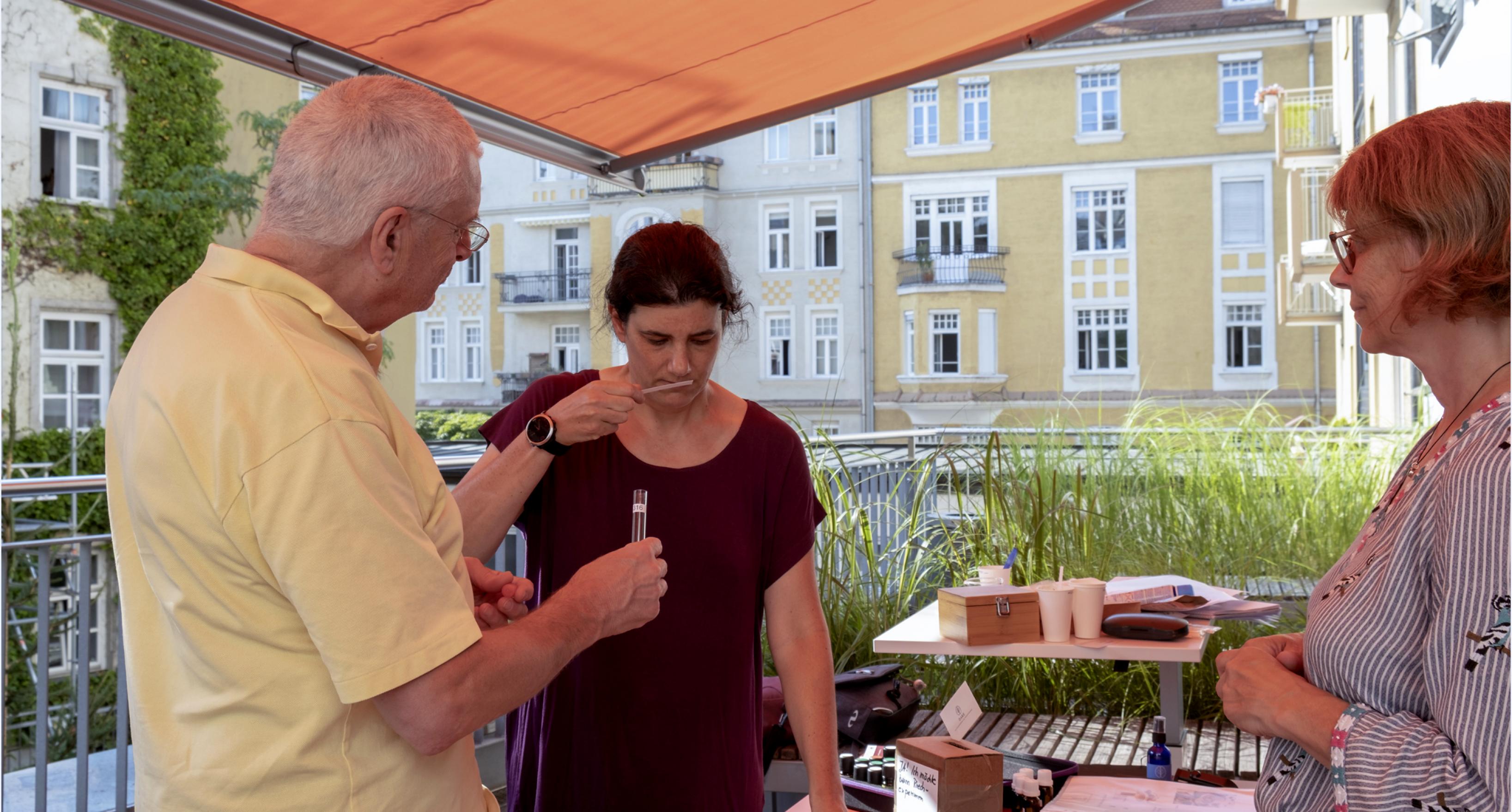 Ein Mann und eine Frau stehen an einem Tisch unter einem Markise im Freien. Er hält ihr ein Röhrchen hin, sie riecht an einem Stäbchen. Die Journalistin Fabienne Hübener steht dahinter.