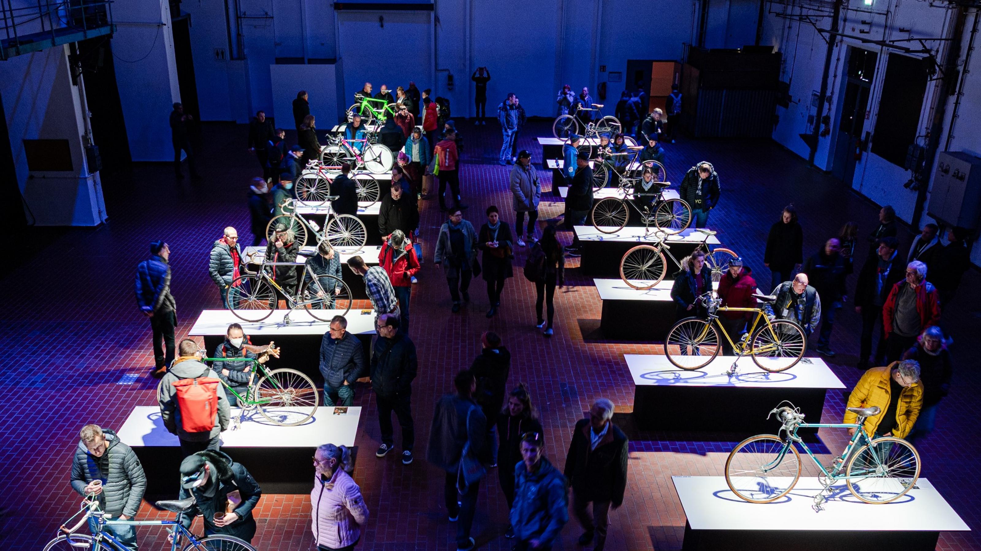 Um längliche, von Schwéinwerfern fokussierte Tische mit aufgestellten Sporträdern scharen sich in der abgedunkelten Industriehalle die Messebesucher.