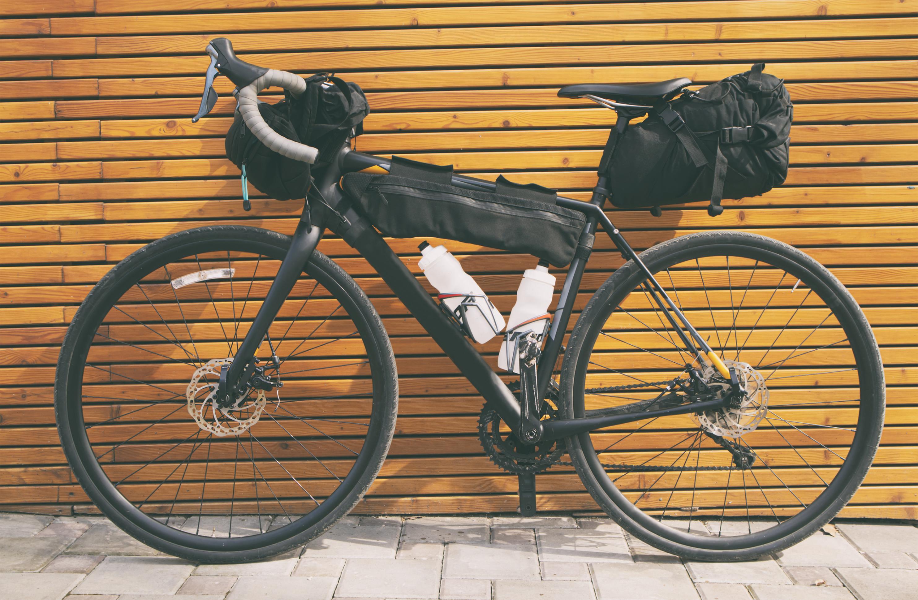 An einer Hilzwand lehnt ein vollbepacktes Fahrrad mit zwei gefüllten Wasserflaschen. Sowohl Lenker als auch Querstang und Sattelstütze tragen Packtaschen.