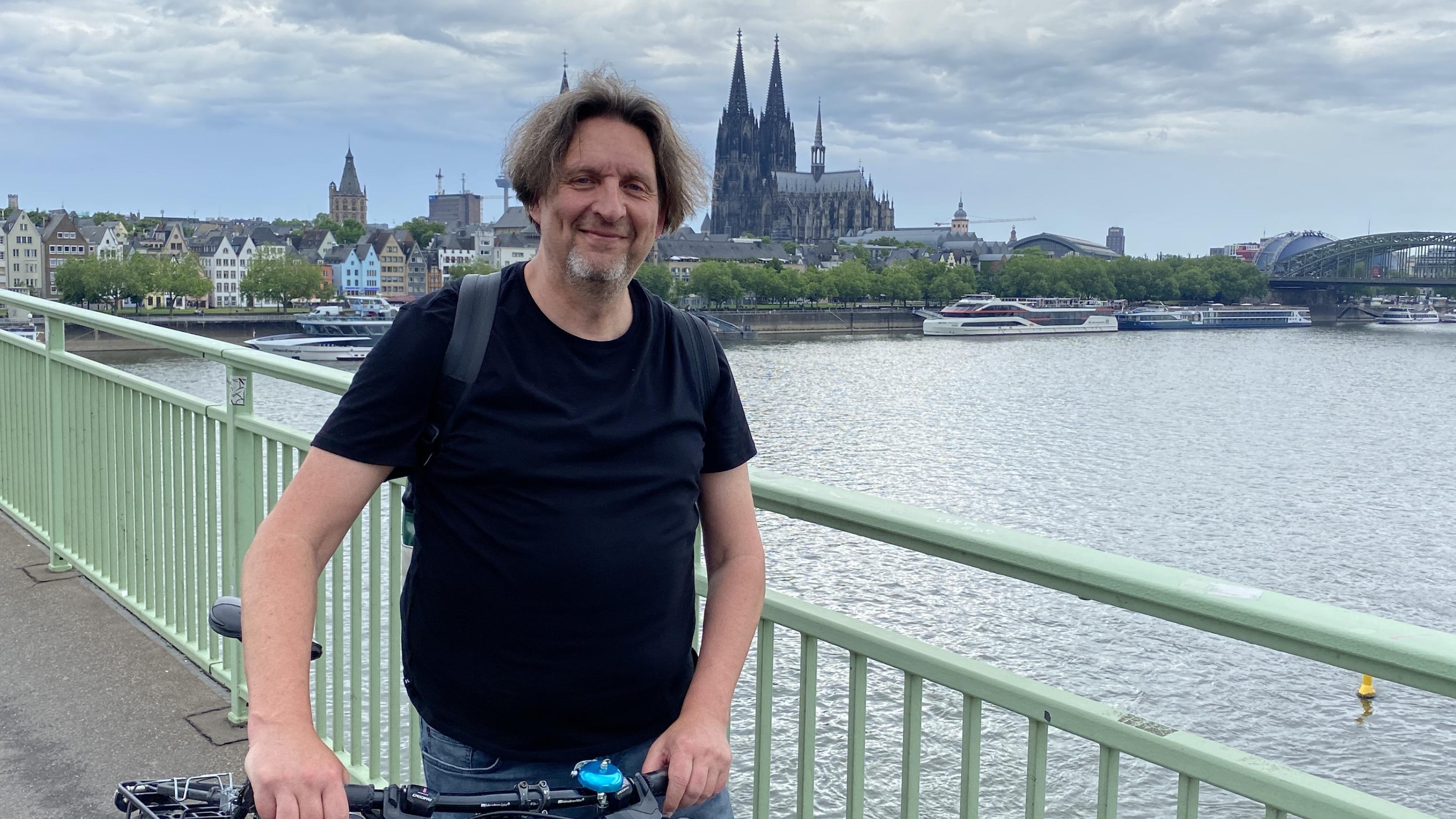 Ein Mann steht mit seinem Fahrrad auf einer Brücke. Im Hintergrund ist der Kölner Dom zu sehen.