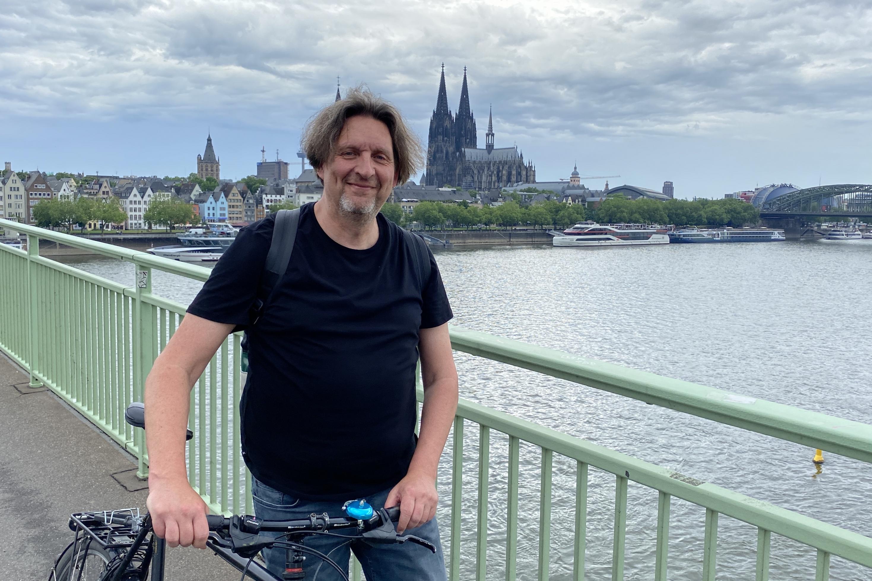 Ein Mann steht mit seinem Fahrrad auf einer Brücke. Im Hintergrund ist der Kölner Dom zu sehen.