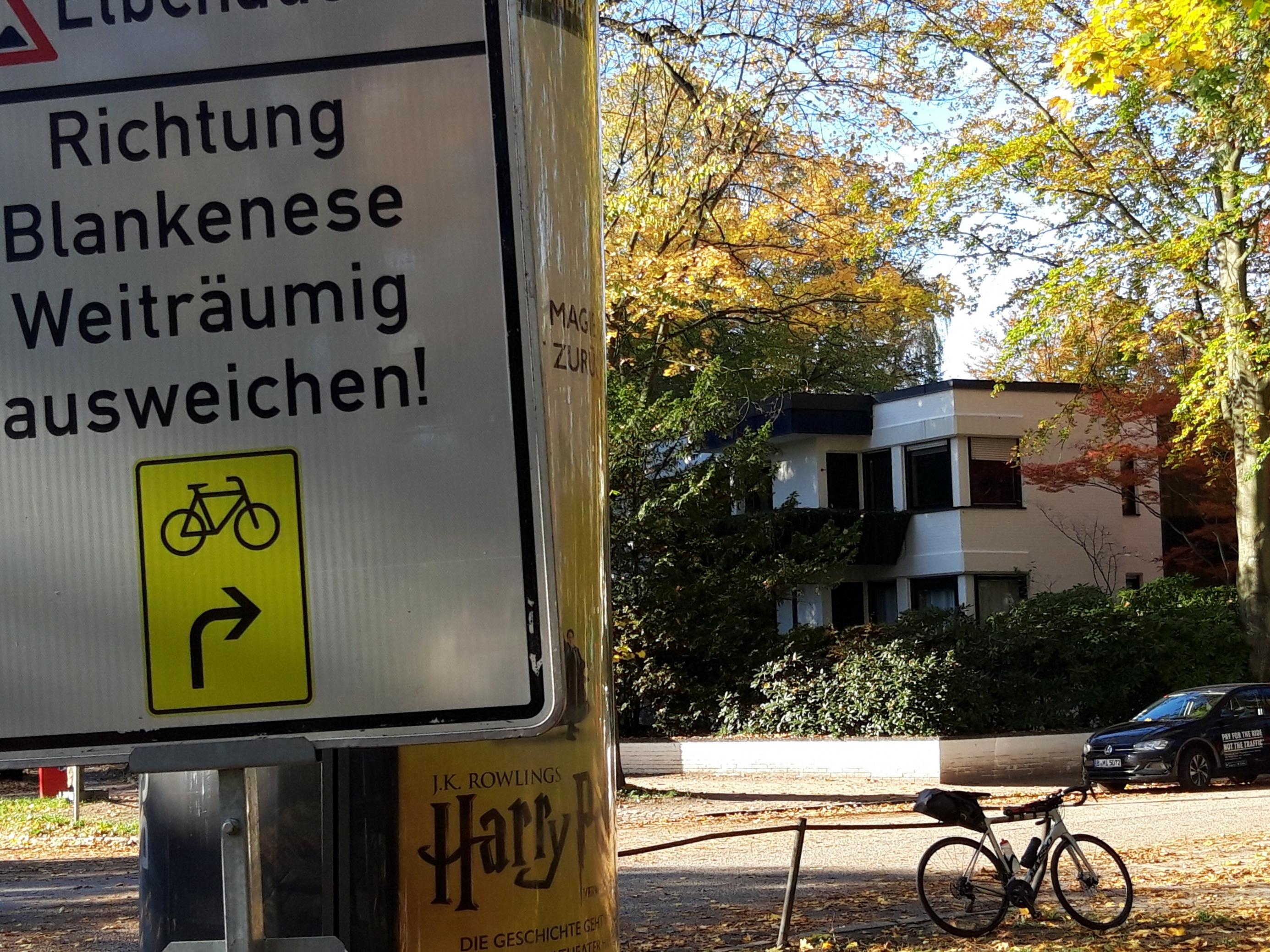 Im Vordergrund ragt die Hinweistafel mit der Inschrift „Richtung Blankenese weiträumig ausweichen!“ Rechts im Hintergrund ist ein weißes Wohnhaus zwischen Bäumen in herbstlicher Tracht zu sehen, davor das Rennrad des Autors.