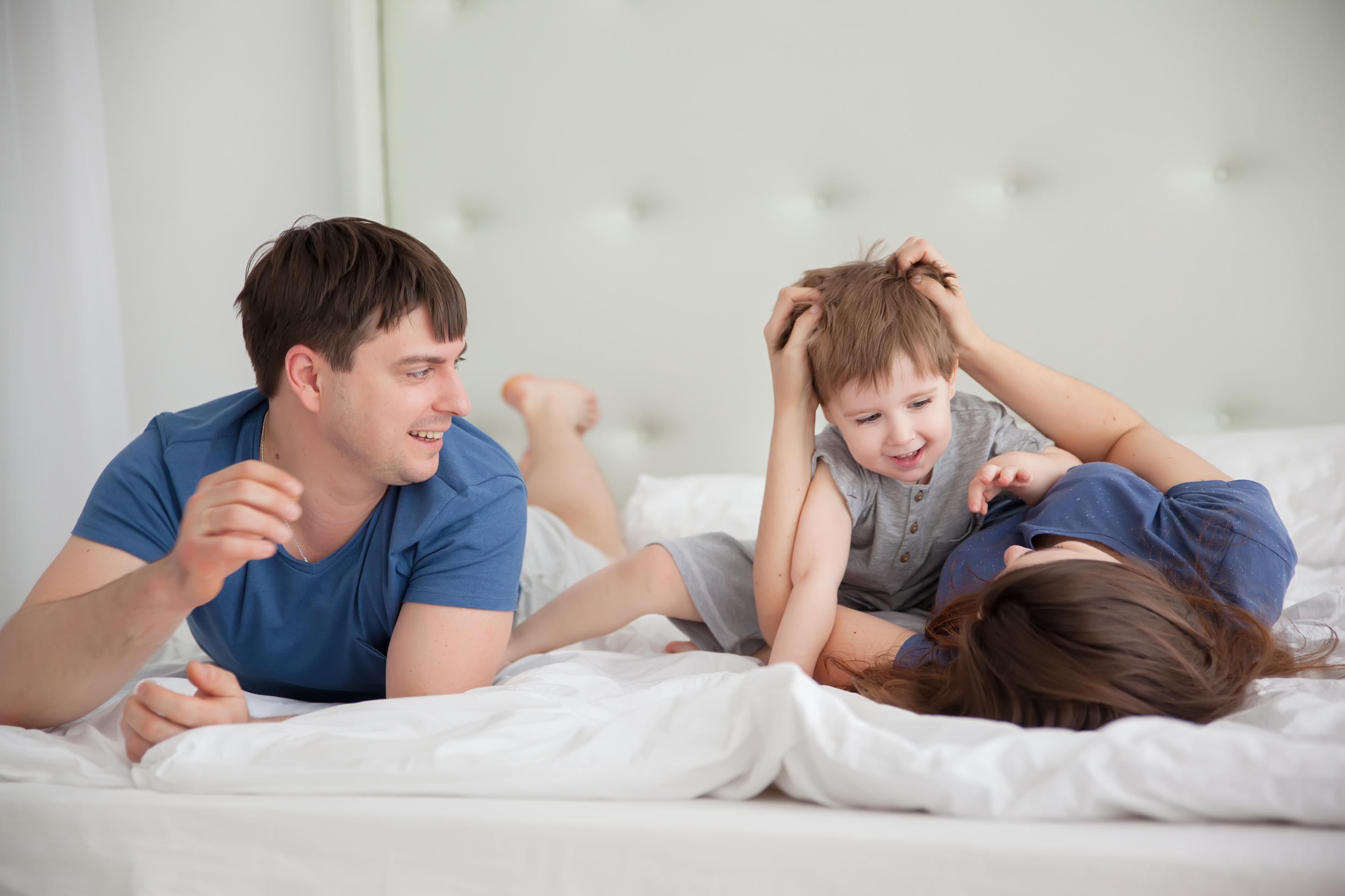 Eine junge Familie – Vater, Mutter, Kind – tobt morgens im Bett. Alle sehen zufrieden und ausgeschlafen aus. Sie haben noch ihre Schlafanzüge an.