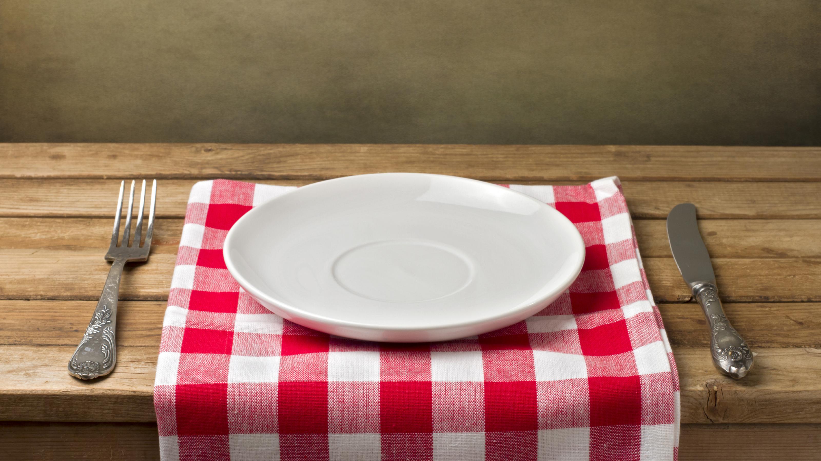 Ein leerer weißer Teller steht auf einem rot-weiß-karierten Set auf einem Holztisch. Messer und Gabel daneben.