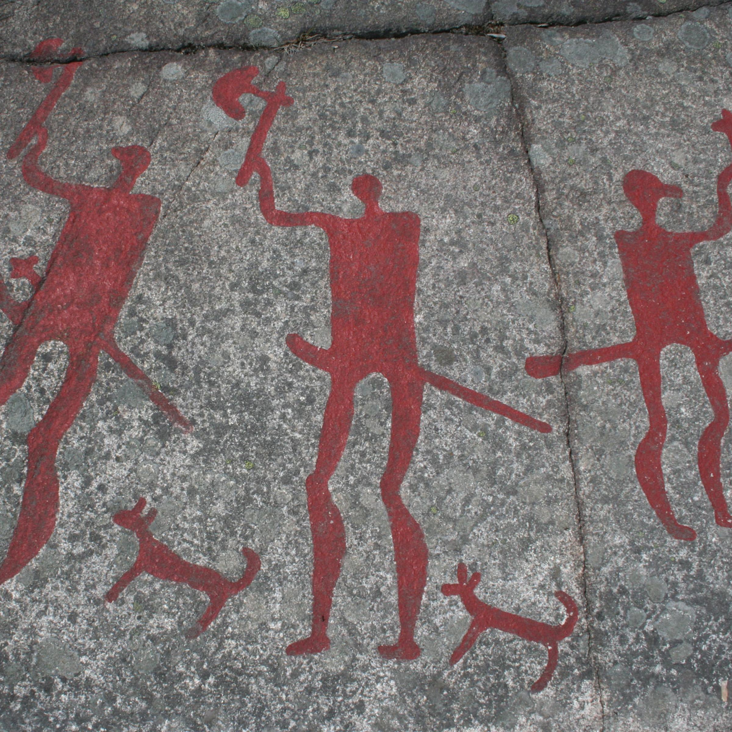 Das Foto zeigt eine Jahrtausende alte Felszeichnung aus der Gemeinde Tanum in Schweden. Prähistorische Menschen haben dort Bilder in den grauen Felsen geritzt und mit leuchtend roten Farben hervorgehoben. Zu sehen sind drei kriegerisch wirkende Figuren sowie drei Hunde.