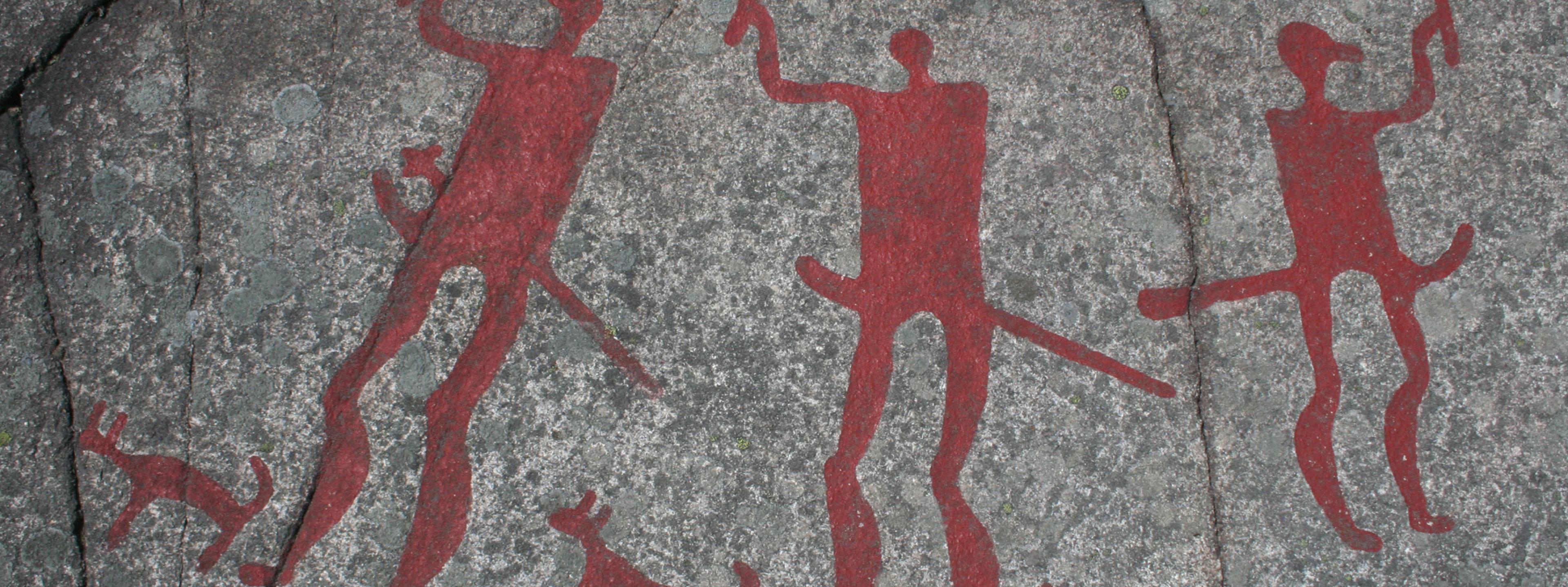 Das Foto zeigt eine Jahrtausende alte Felszeichnung aus der Gemeinde Tanum in Schweden. Prähistorische Menschen haben dort Bilder in den grauen Felsen geritzt und mit leuchtend roten Farben hervorgehoben. Zu sehen sind drei kriegerisch wirkende Figuren sowie drei Hunde.