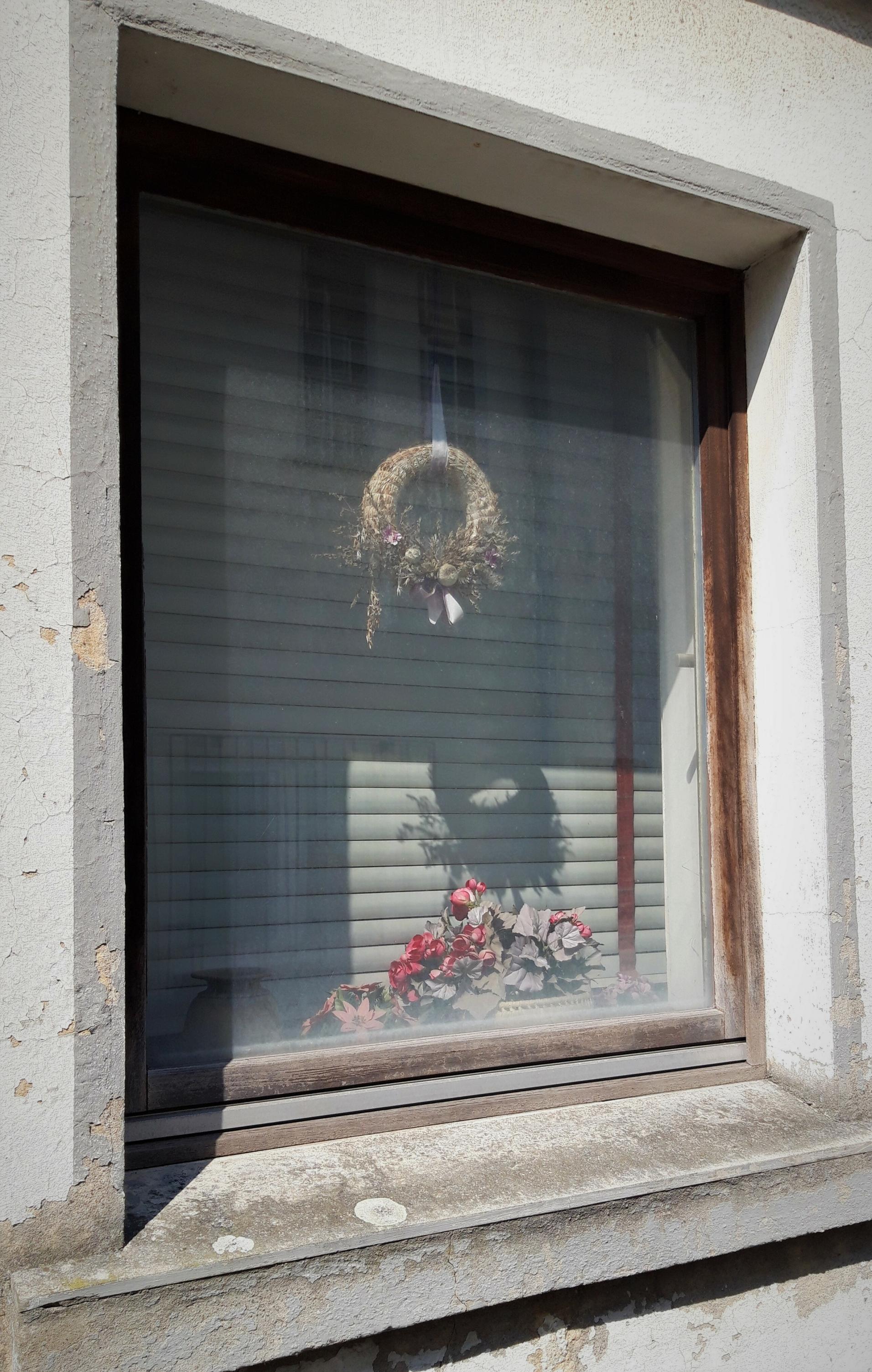 In einer schäbig-weißen Hauswand tut sich eine Fensteröffnung auf. Zwischen Glas und heruntergelassener Jalousie hängt ein staubiger Flechtkranz; darunter steht ein kleines Bouquet aus Plastikblumen.