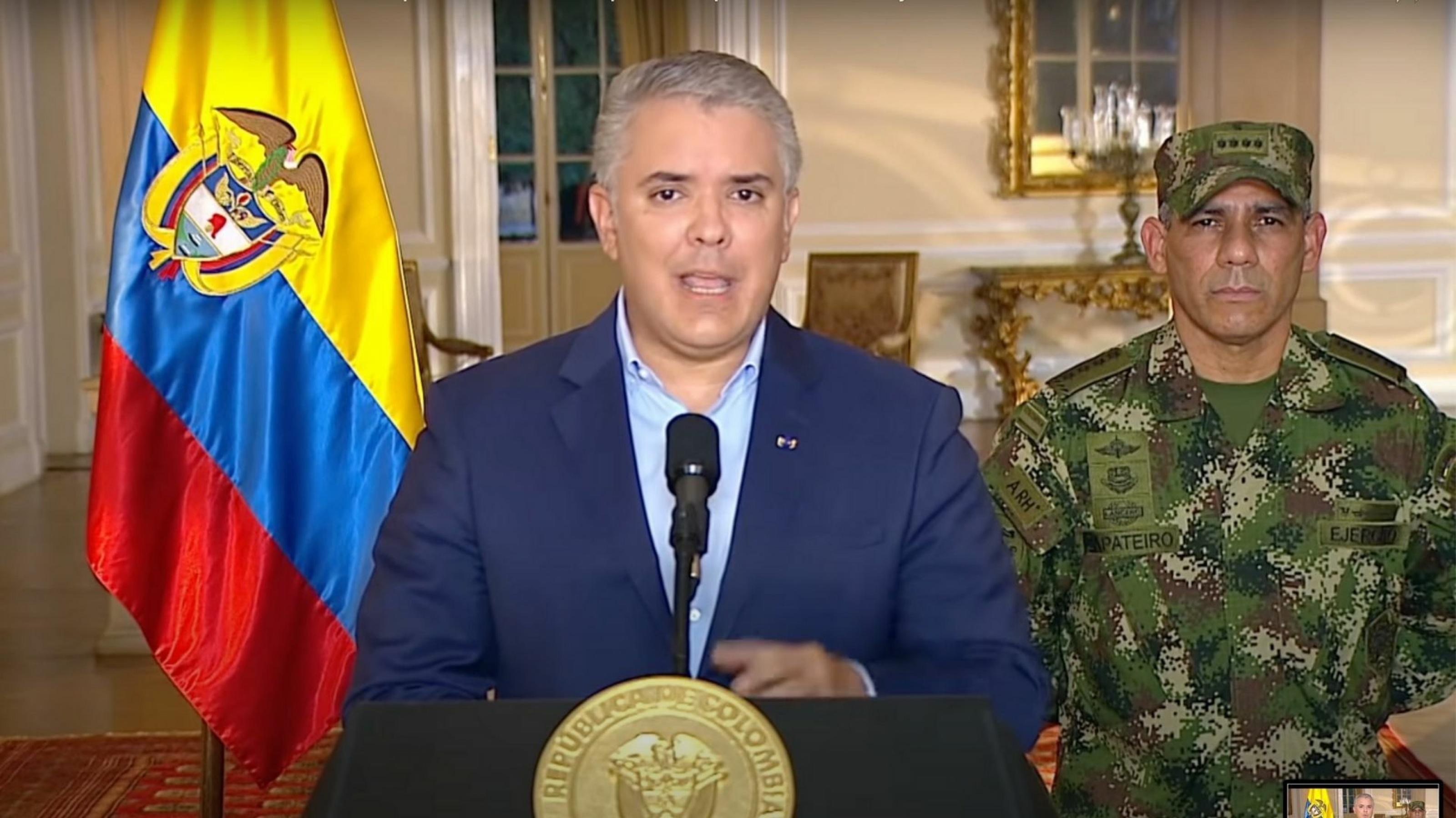 Ein Mann mit jungem Gesicht und grauen Haaren steht am Redepult im Präsidentschaftspalast. Er trägt einen Anzug mit Hemd ohne Krawatte. Hinter ihm stehen die kolumbianische Flagge und ein General in Uniform mit ernstem Gesicht.