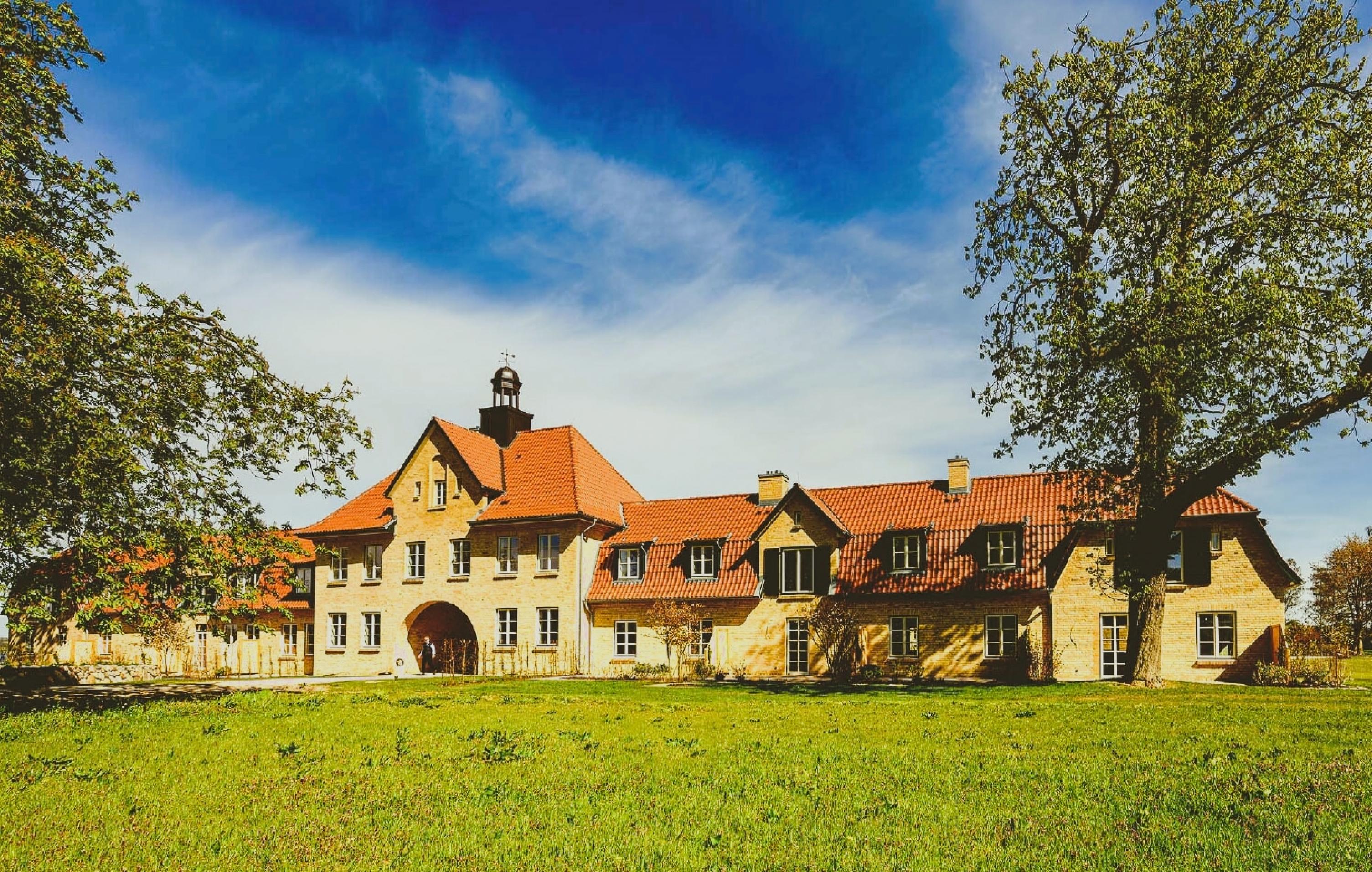 Stattlicher Torhaus-Komplex des Gutshofs, abgelichtet hinter einer üppigen Wiese und zwischen alten knorrigen Laubbäumen.