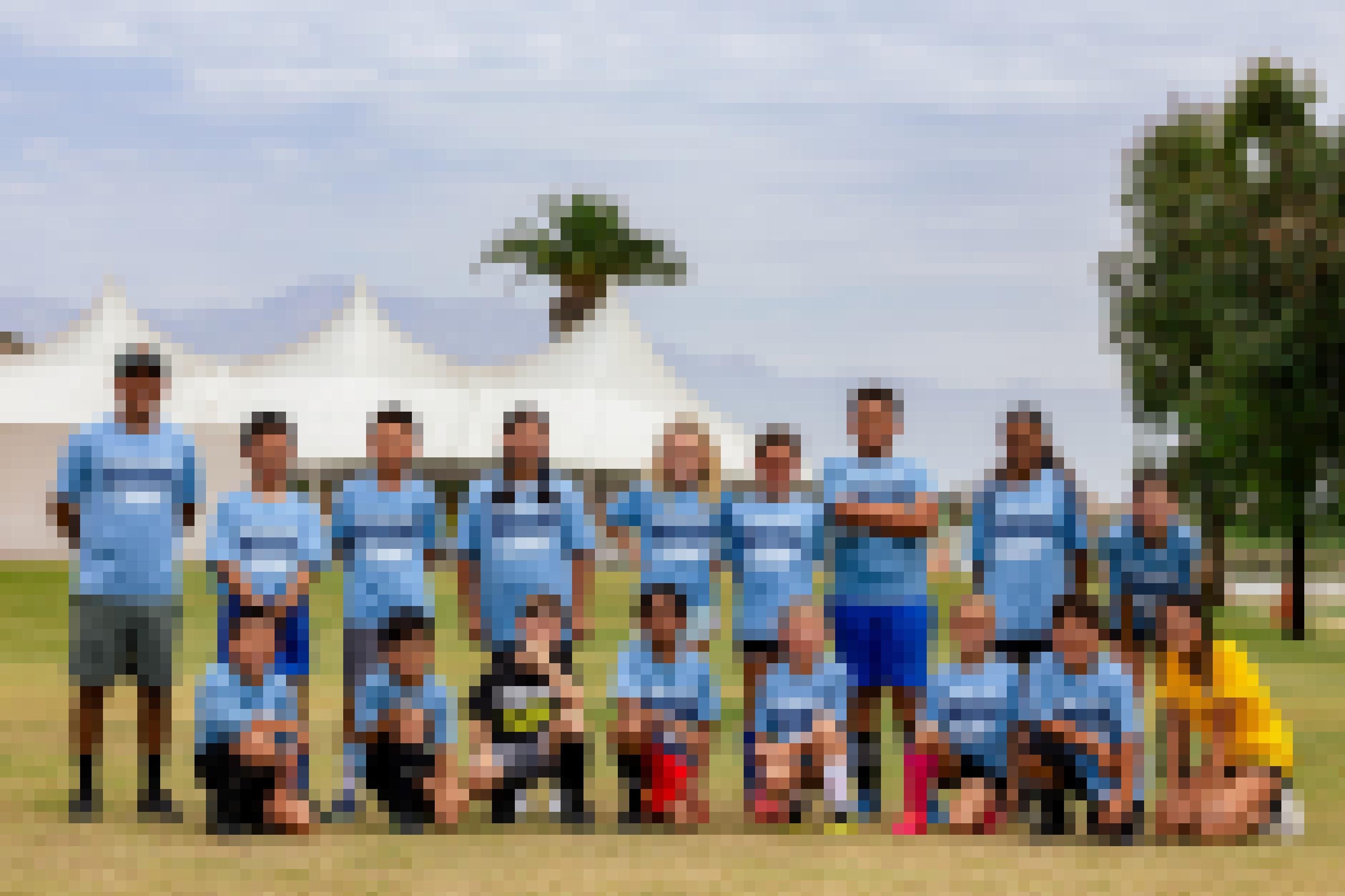 Jürgen und Mädchen einer gemischten Fußballmannschaft posieren mit ihrem Trainer in hellblauen Trikots auf dem Feld. Im Hintergrund ein Zelt, Palmen und Berge.