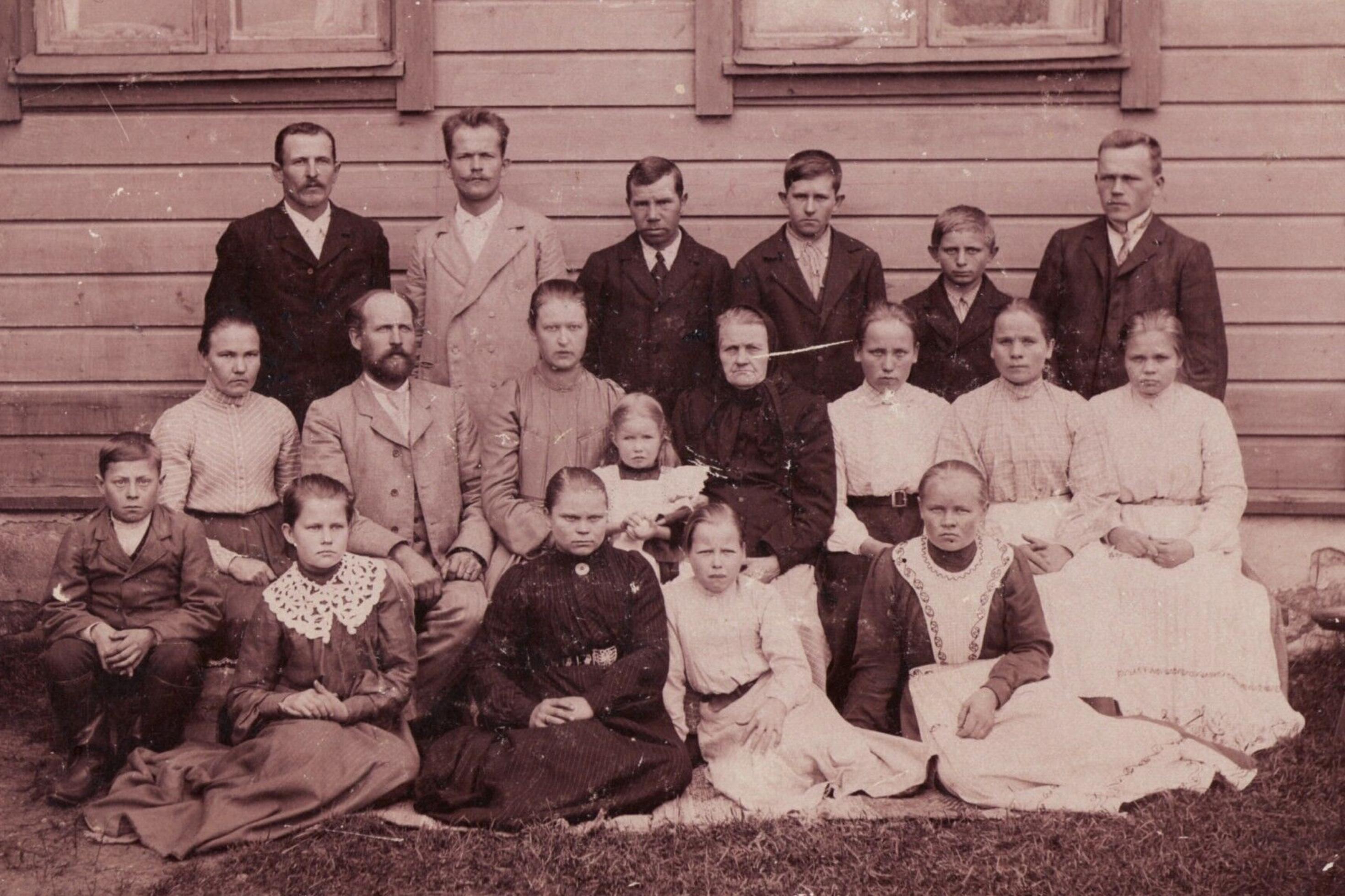 Das historische Schwarzweißfoto zeigt 19 Mitglieder einer finnischen Familie, die vor der Bretterwand eines Holzhauses posieren. Es sind Frauen, Männer und Kinder unterschiedlichen Alters, die mit ernstem Blick in die Kamera schauen, alle herausgeputzt mit Blusen und Röcken bzw. Kleidern oder mit Anzügen.