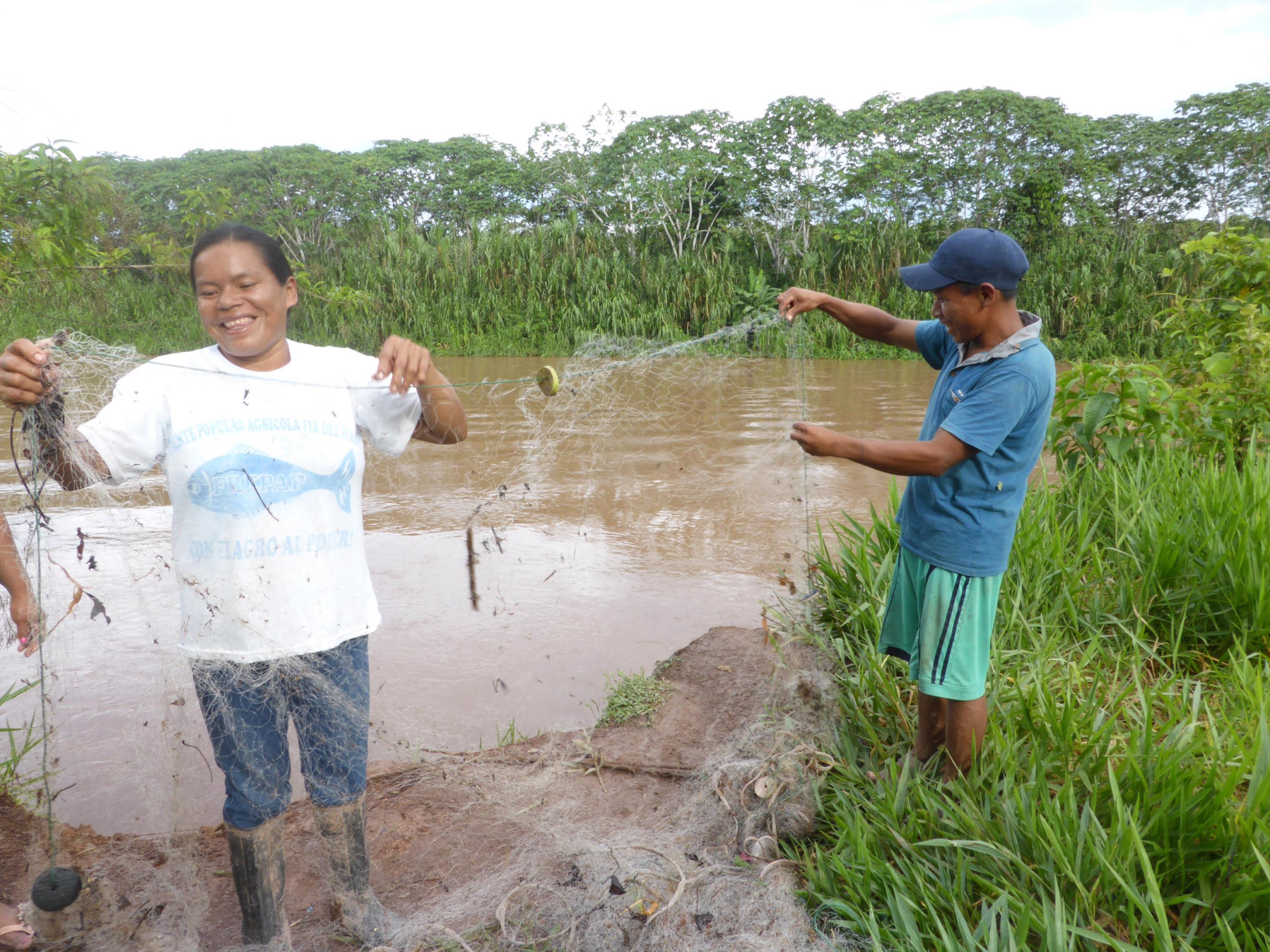 Die Fischerin steht in Gummistiefeln am matschigen Ufer des Amazonas und hält ihr Netz hoch, in dem kleine Holzstücke hängen. Ihr Mann hilft ihr und hält das andere Ende.
