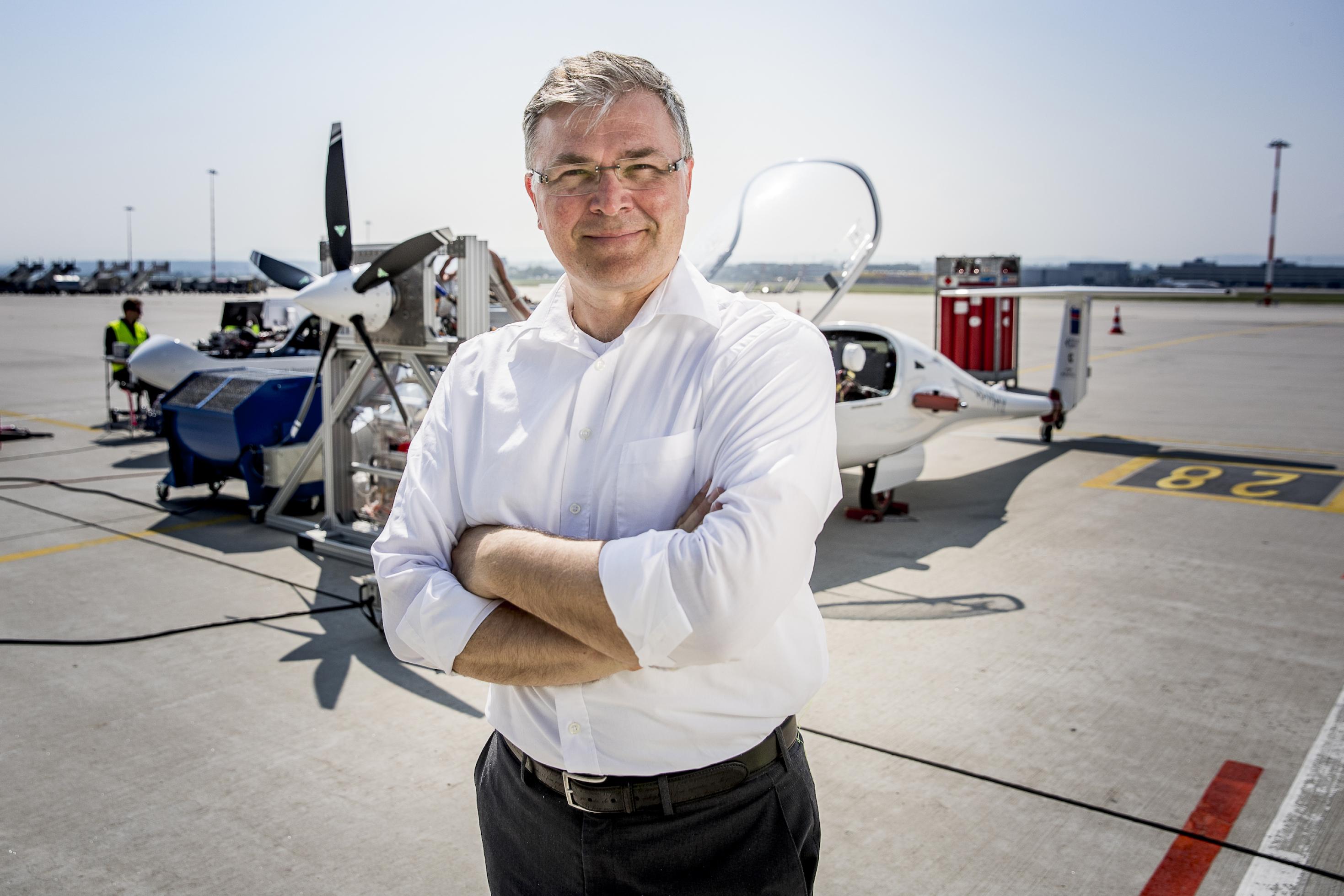 Ein mittelalter Mann mit silberfarbenem Haar steht mit weißem Hemd und verschränkten Armen vor einem zweimotorigen Flugzeug bei Sonnenschein.