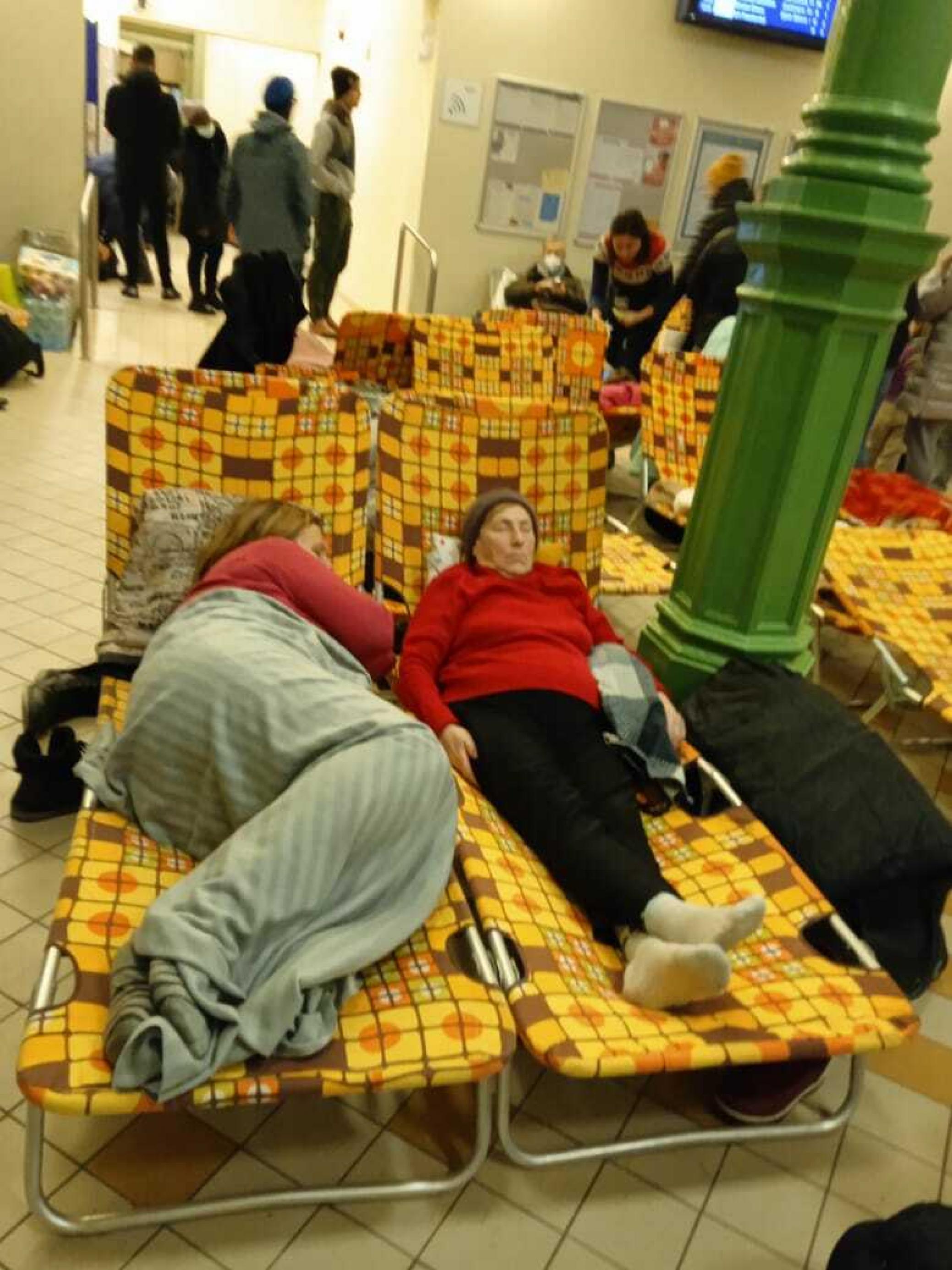 Zwei Frauen ruhen sich nach der Flucht auf gelben Liegen aus, dahinter stehen weitere Menschen in dem polnischen Bahnhofsgebäude