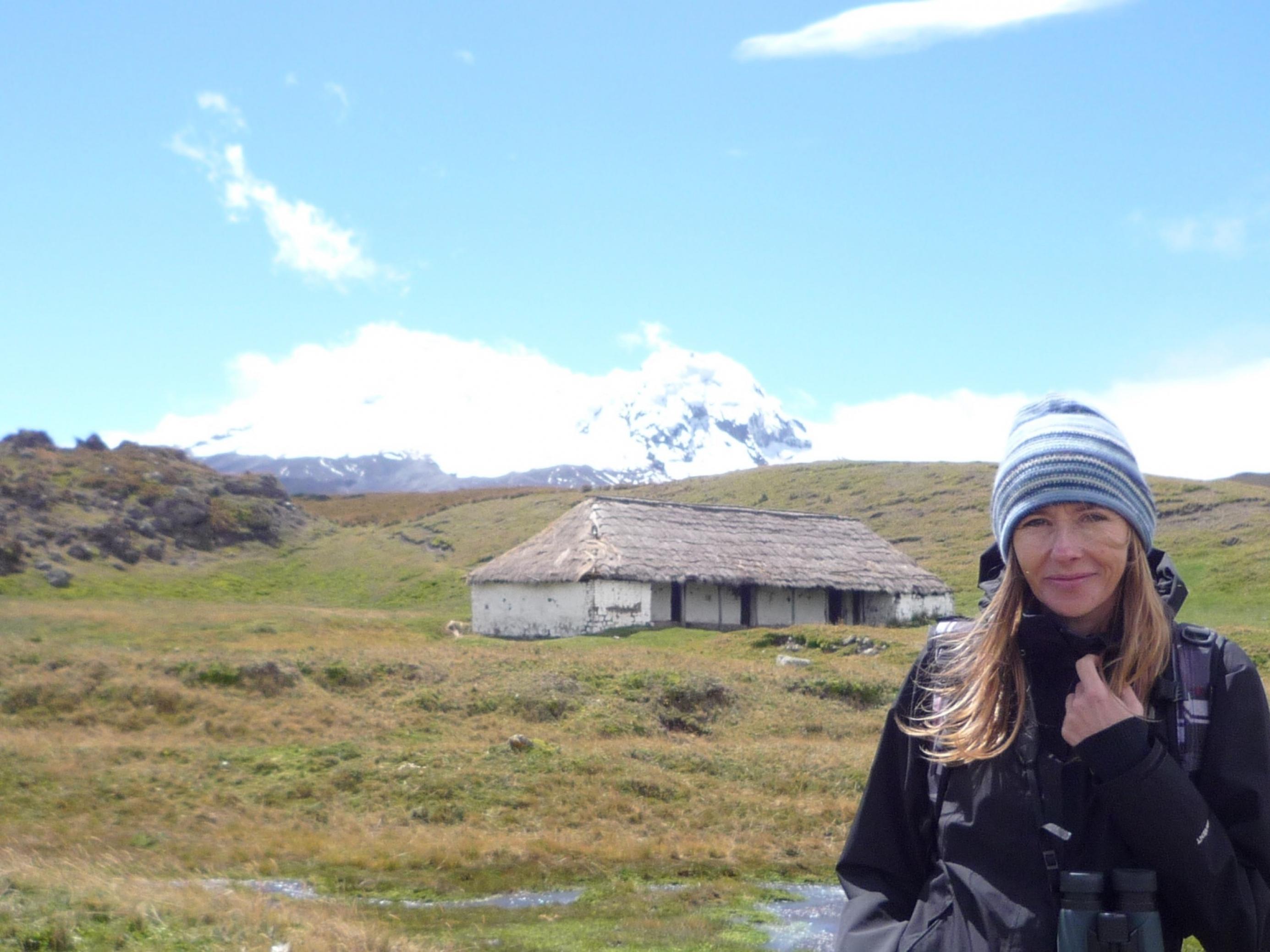 Aufnahme von Andrea Wulf. Sie trägt eine Mütze und steht vor einer hügeligen Landschaft mit einem alten Gebäude. Im Hintergrund sieht man einen schneebedeckten Berg.