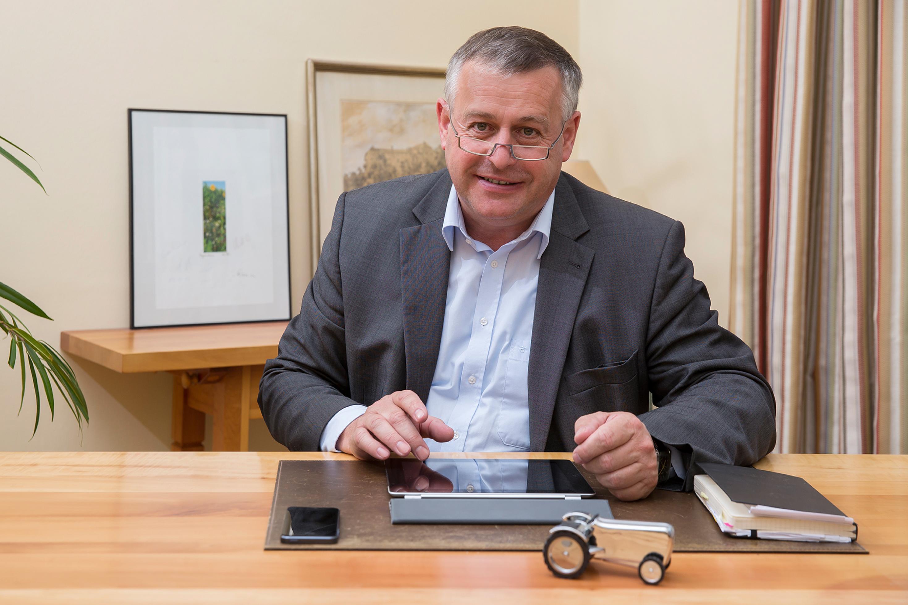 Foto von Walter Heidl an seinem Schreibtisch. Vor ihm liegt ein Tablet, ein Smartphone und ein Buch.