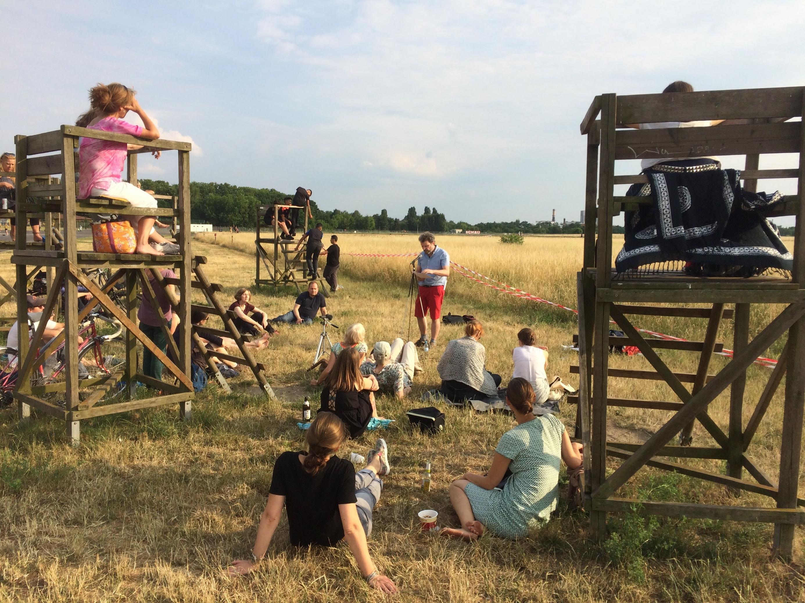 Ein Gruppe Menschen haben sich auf einer Wiese versammelt. Einige sitzen auf dem Boden, andere auf Holzgerüsten. Alle gucken in die Richtung eines Mannes, der vor ihnen an einem Mikrofon steht.