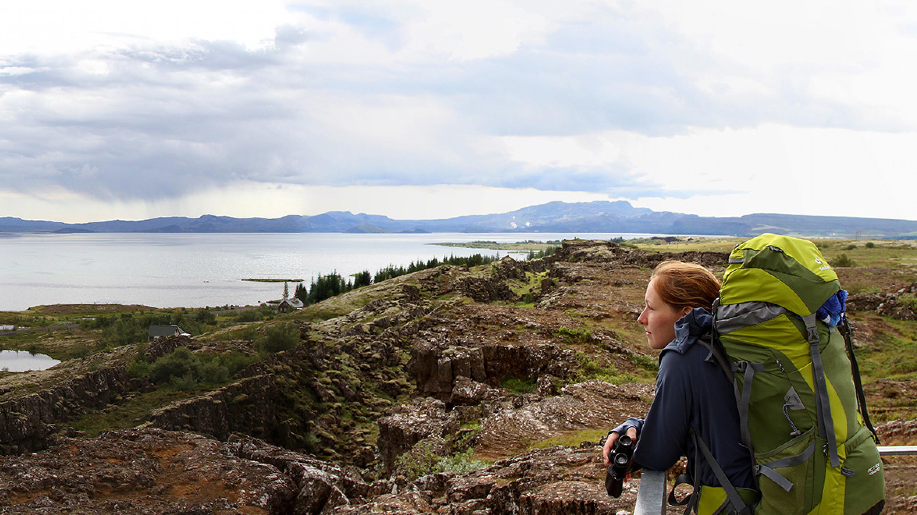 Lisa Pannek im Þingvellir-Nationalpark in Südwestisland. Sie trägt einen großen Wanderrucksack und steht mit einem Fernglas in der Hand vor einer felsigen Landschaft. Im Hintergrund sieht man Wasser und Berge.