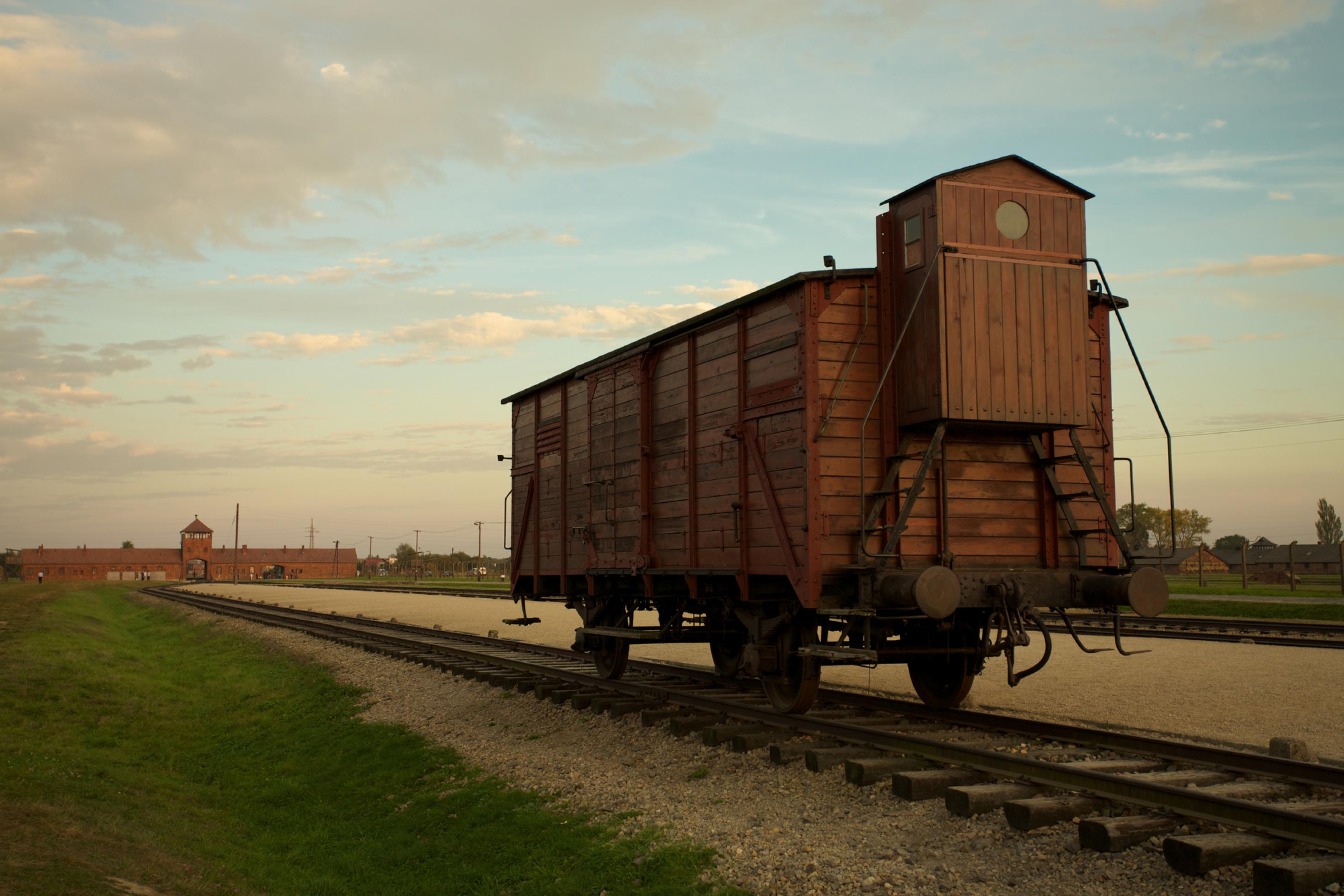 Ein einzelner, geschlossener Waggon auf Schienen. Im Hintergrund sind man das Konzentrationslager Auschwitz-Birkenau