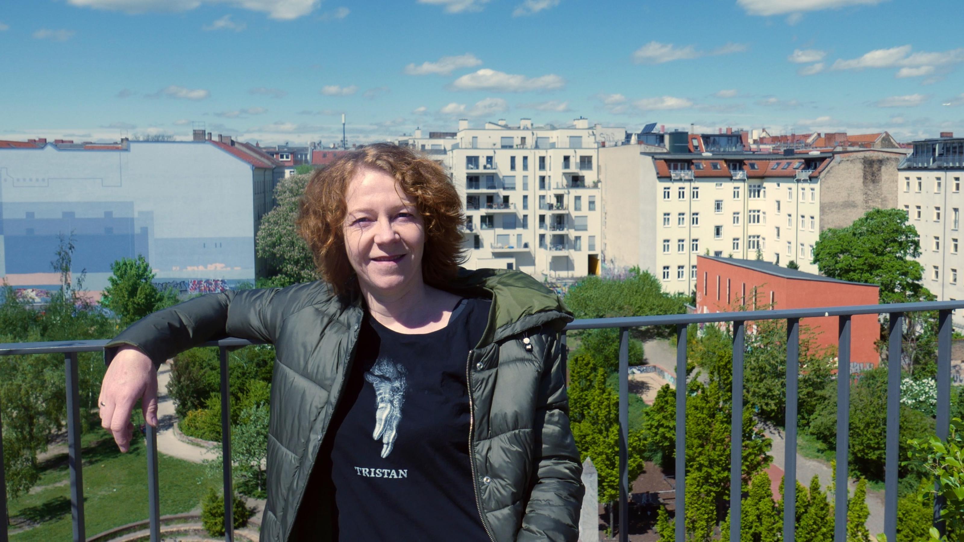 Christiane Habermalz lehnt an einem blauem Geländer. Im Hintergrund kann man einen Park und mehrstöckige Wohnhäuser erkennen.