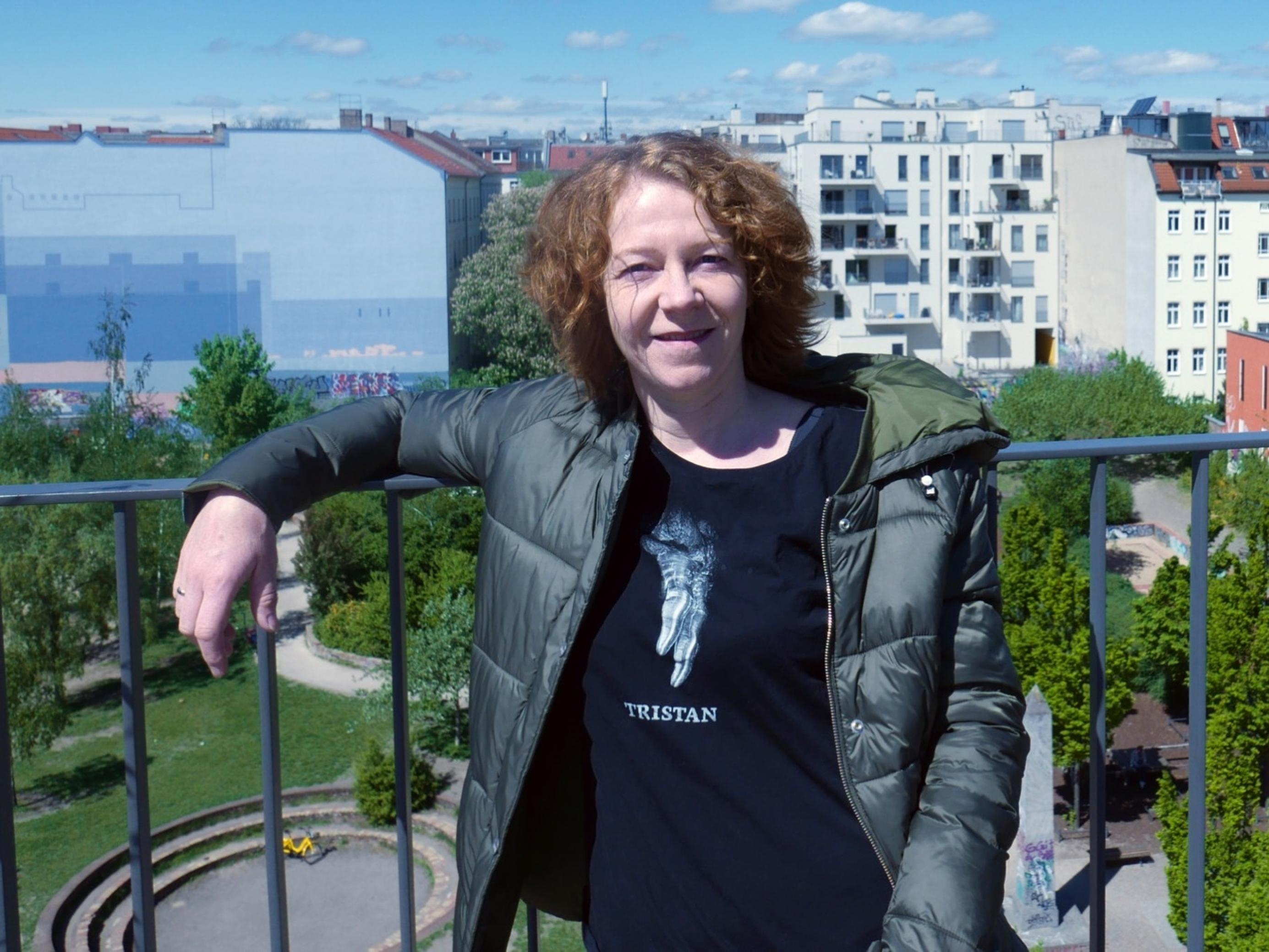 Christiane Habermalz lehnt an einem blauen Geländer. Im Hintergrund erkennt man einen Park und mehrstöckige Wohnhäuser.