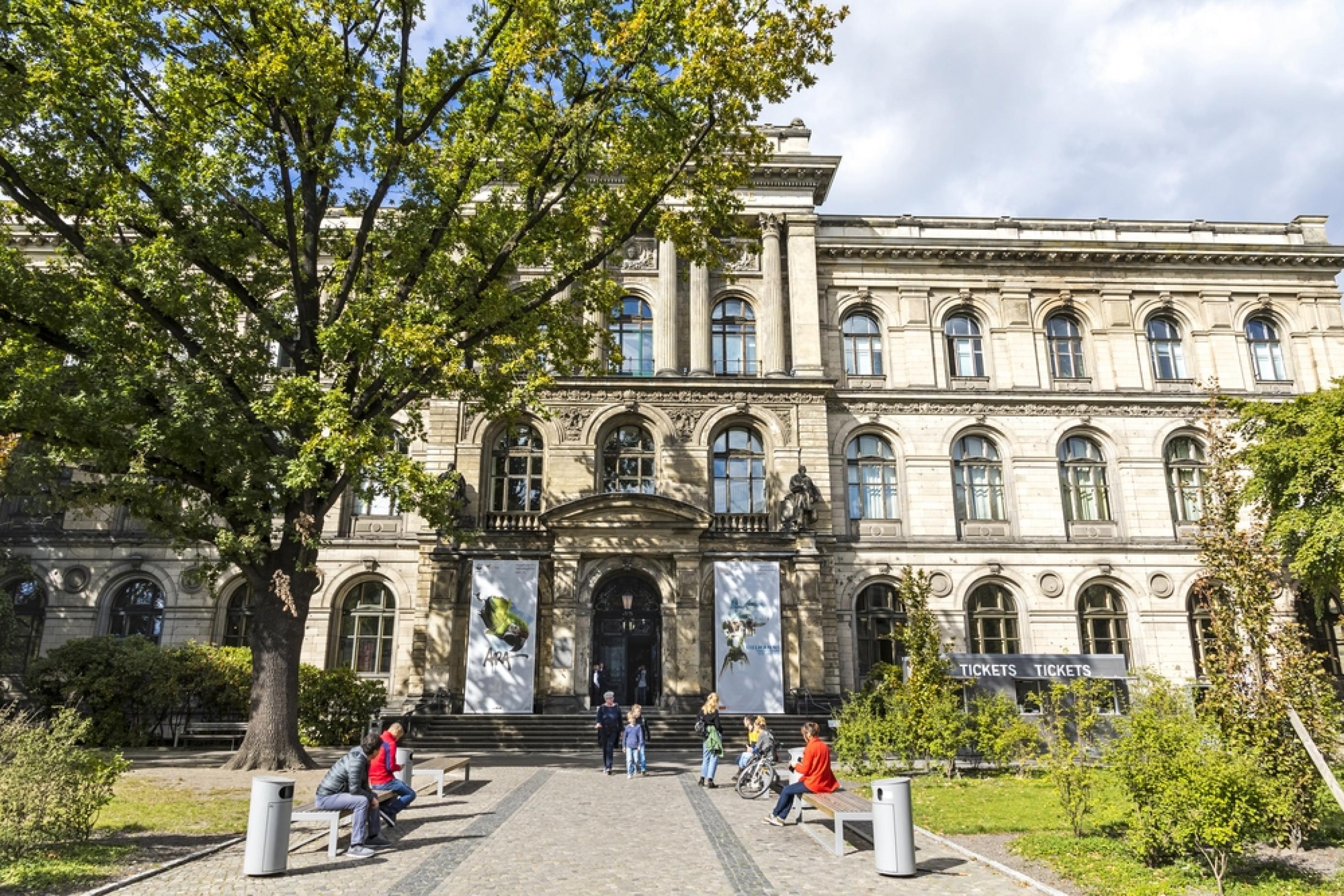 Das Museum für Naturkunde in Berlin. Ein helles, dreistöckiges Gebäude.Vor dem Eingang sind ein paar Bänke auf denen Menschen sitzen.