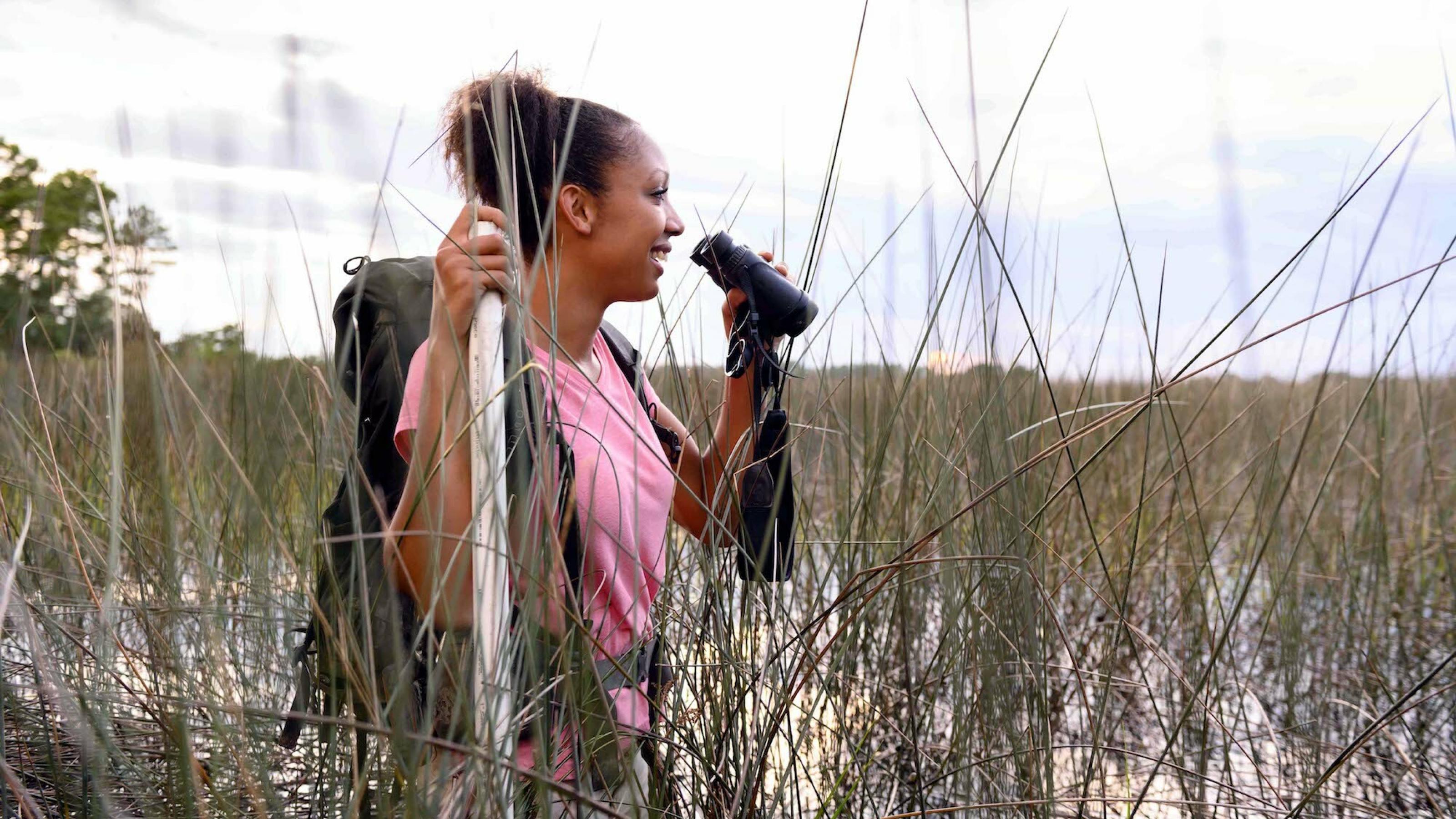 Zu sehen ist die junge Ornithologin Corina Newsome, die mit einem Fernglas in der Hand in einem Sumpf bis zur Hüfte im Wasser steht.