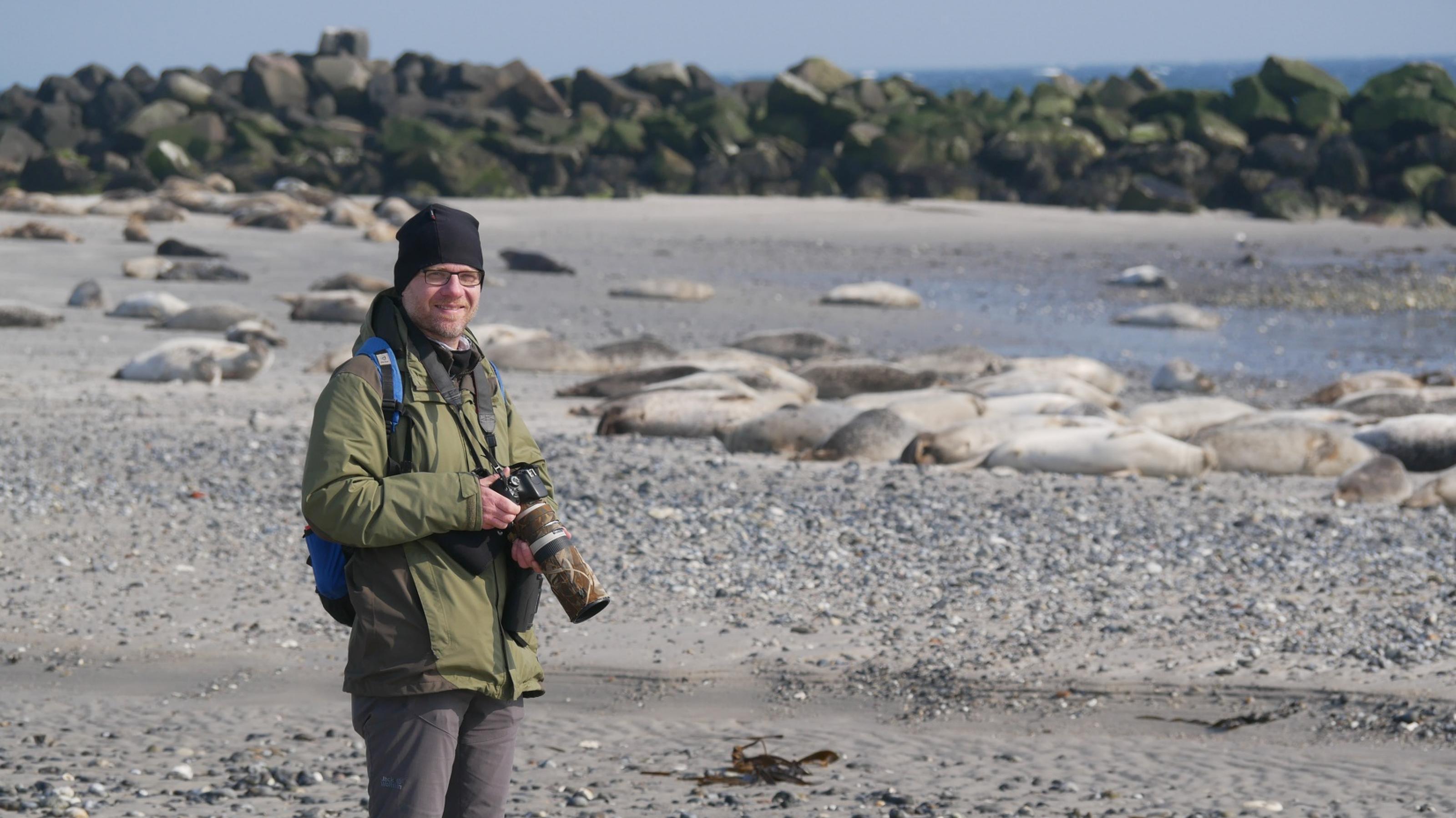 Thomas Gerl steht mit seiner Kamera in der Hand an einem Strand.