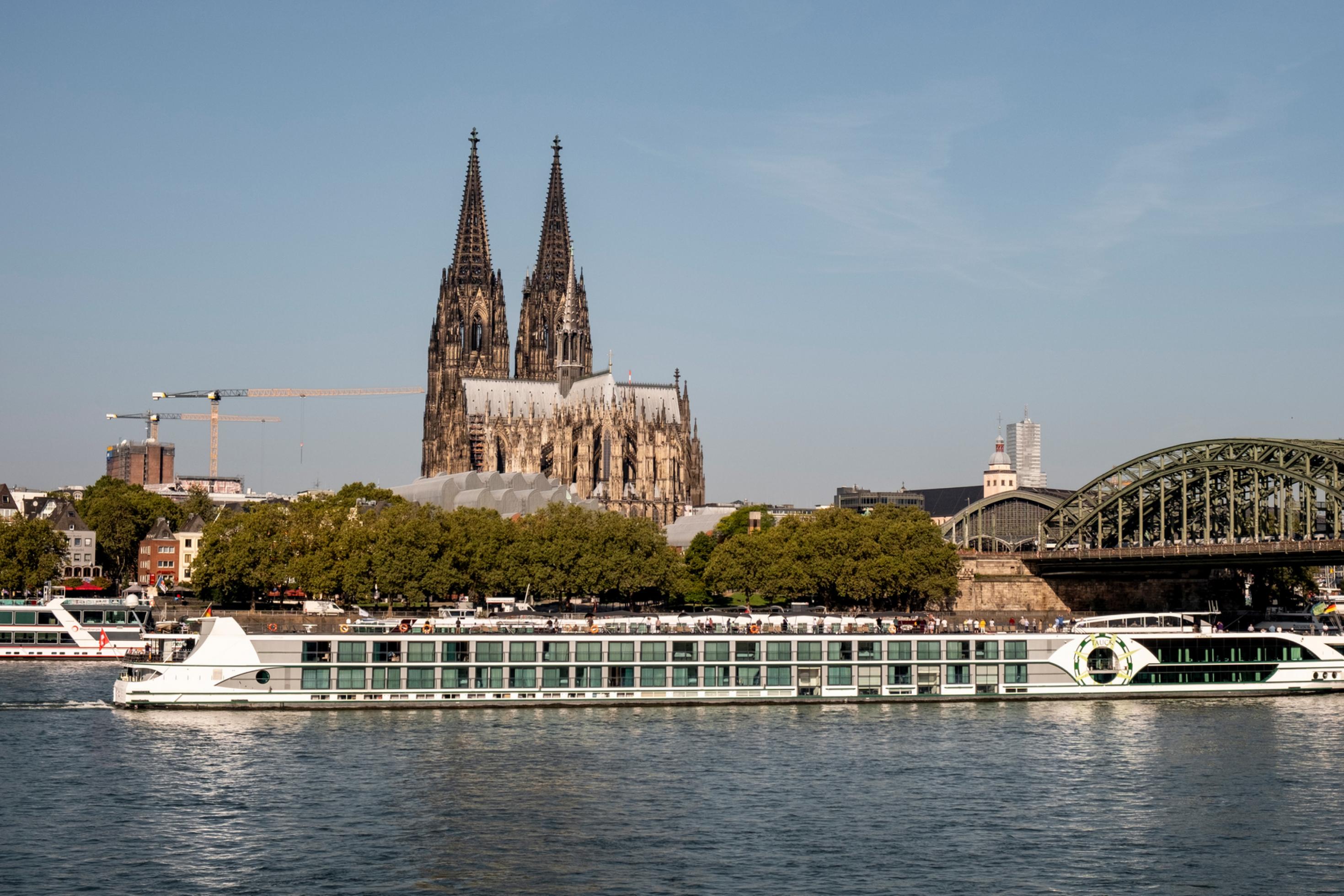 Blick auf den Rhein mit einem Kreuzfahrtschiff darauf, im Hintergrund der Dom und eine Eisenbahnbrücke