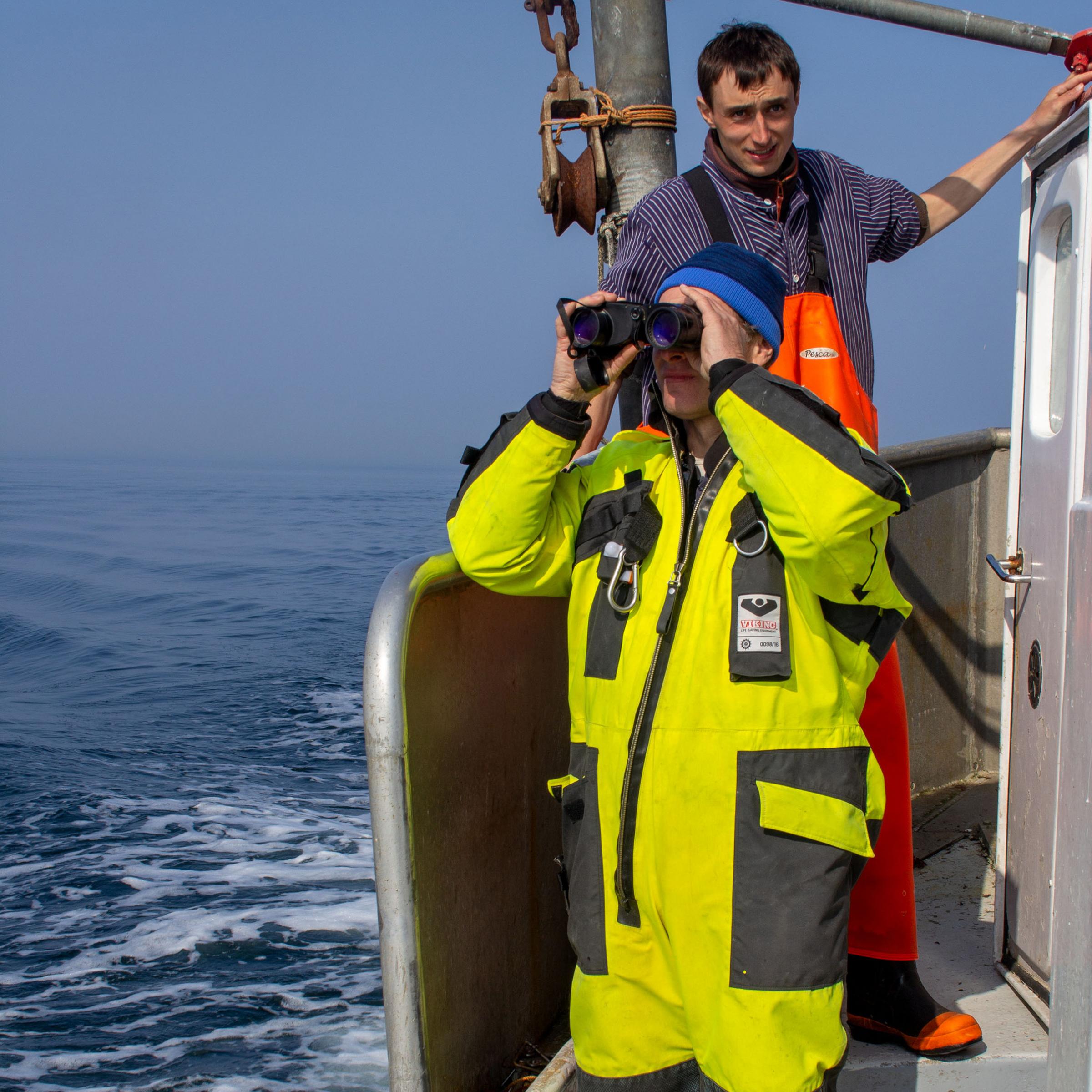 Zwei Männer in Ölzeug auf einem Fischerboot. Einer schaut durch ein Fernglas.