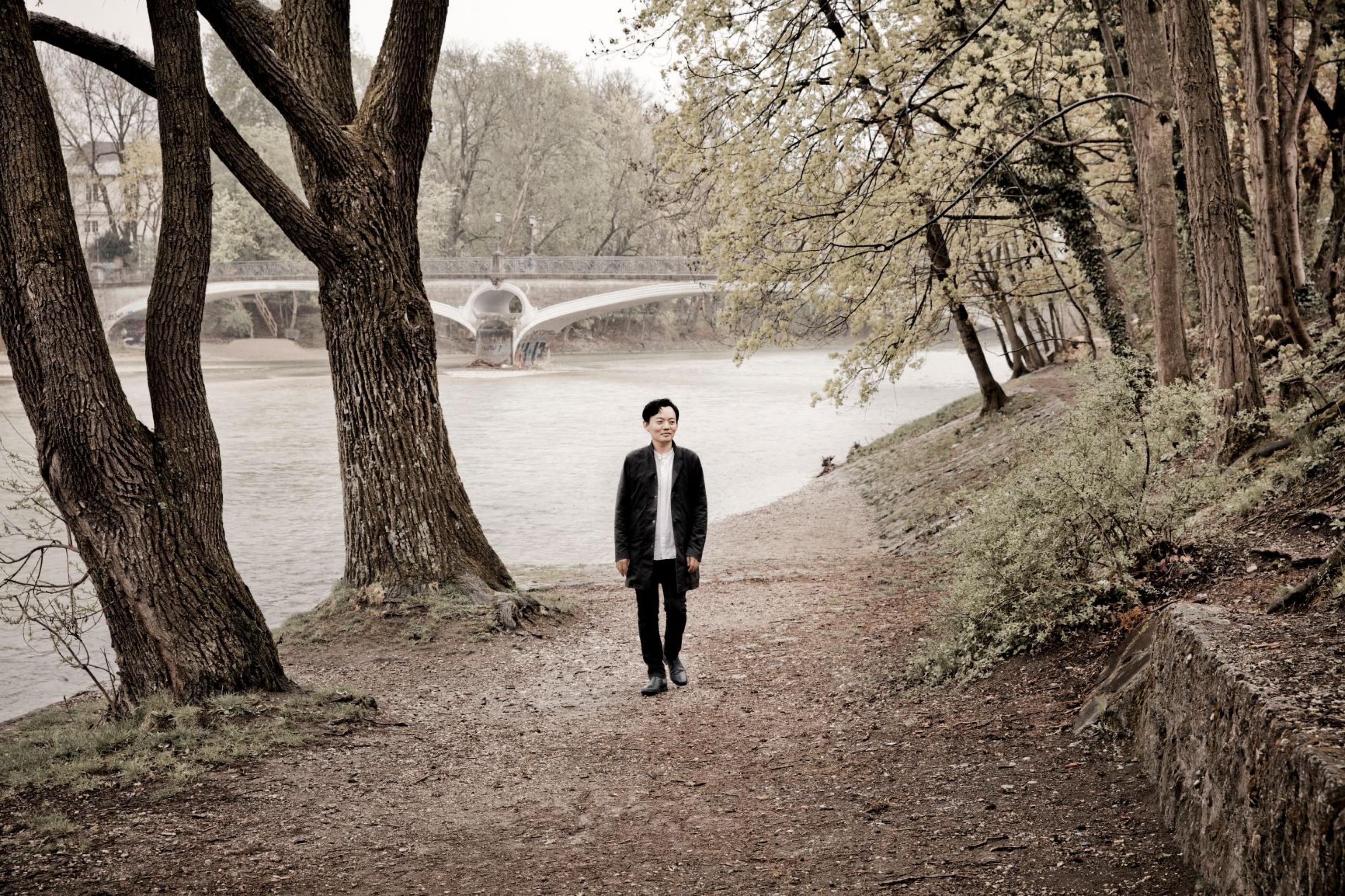Der Pianist William Youn läuft im weißen Hemd und schwarzer Jacke am Ufer eines Flusses entlang.