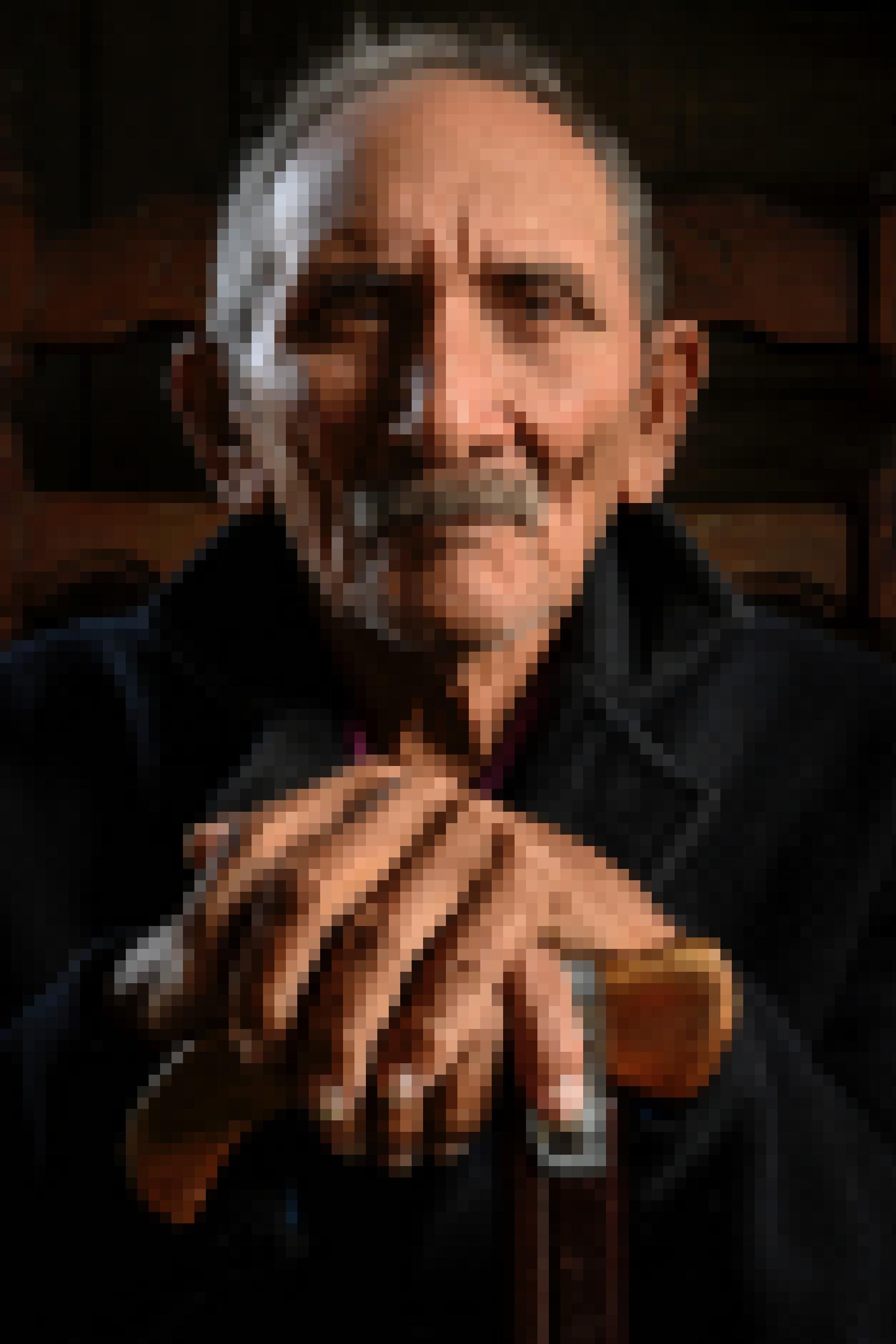 Alter Mann mit Schnurrbart und einer schwarzen Jacke, deren Kragen aufgestellt ist, blickt fest in die Kamera, seine Hände ruhen auf einen Gehstock