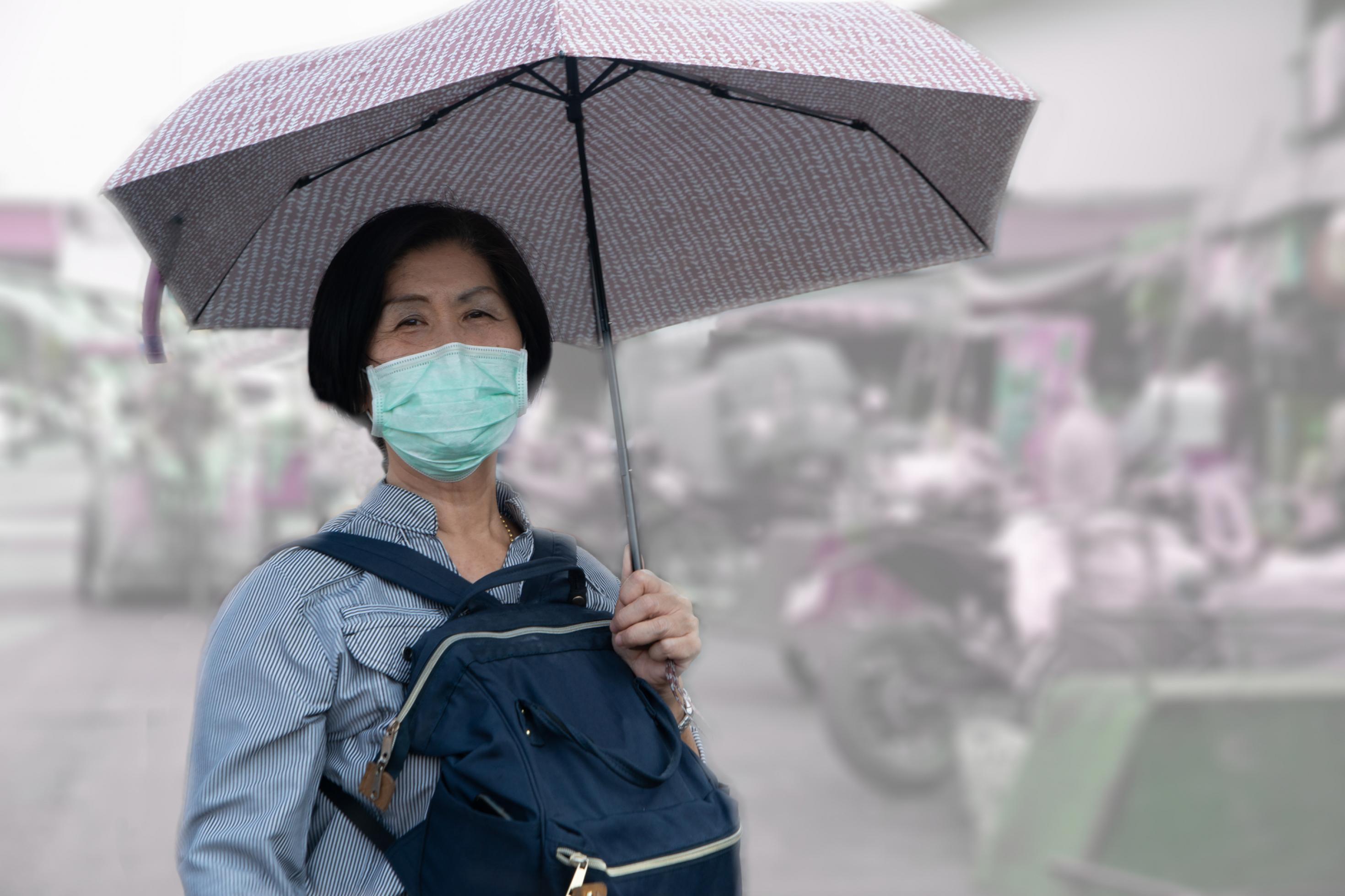 Eine ältere Frau mit Regenschirm und Schutzmaske vor dem Gesicht auf einem asiatischen Markt.