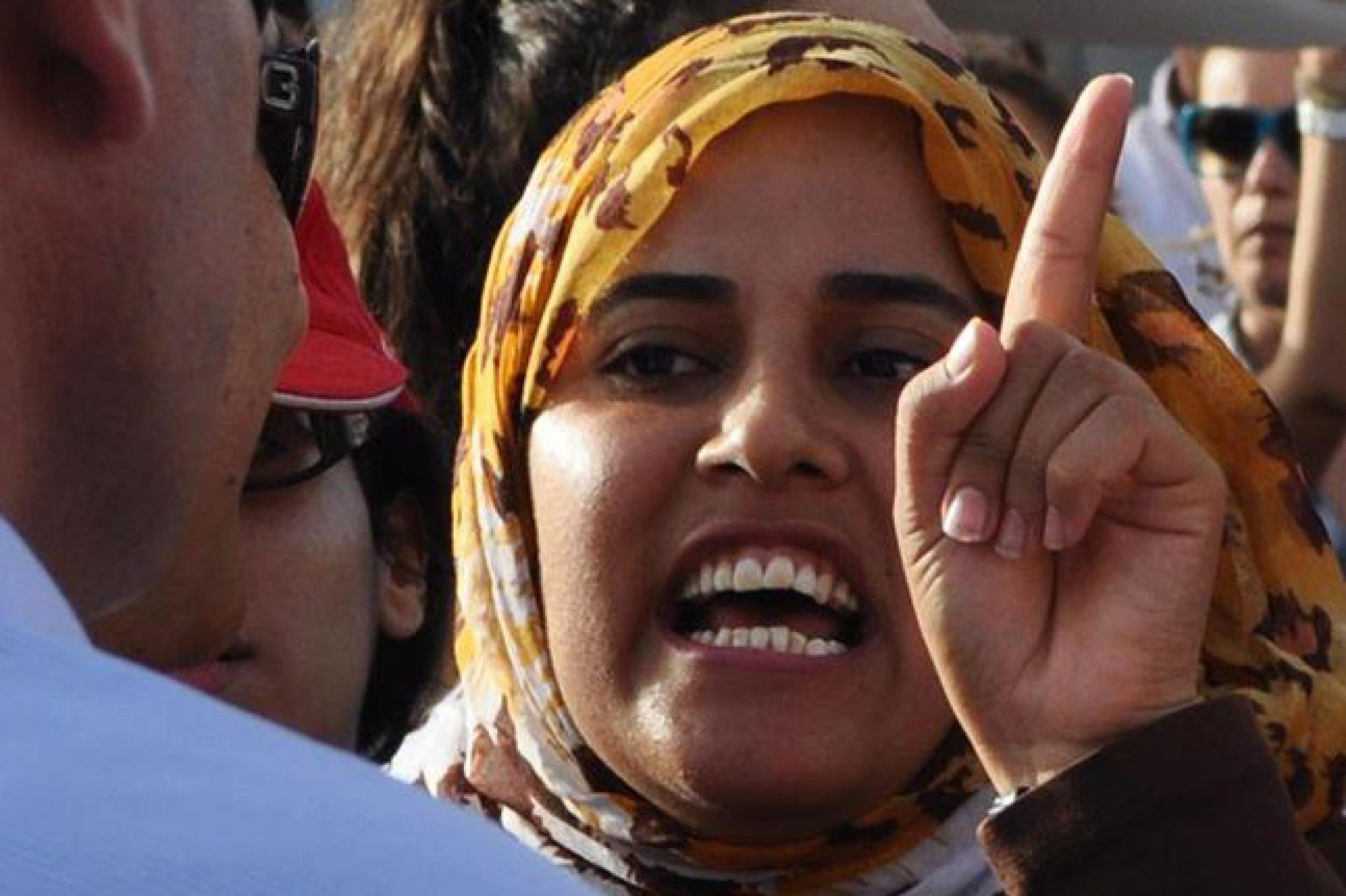 Frau mit gelb-braun gemustertem Kopftuch, die mit erhobenem Zeigefinger auf einer Demonstration in Tunis einem Polizisten etwas zuruft.
