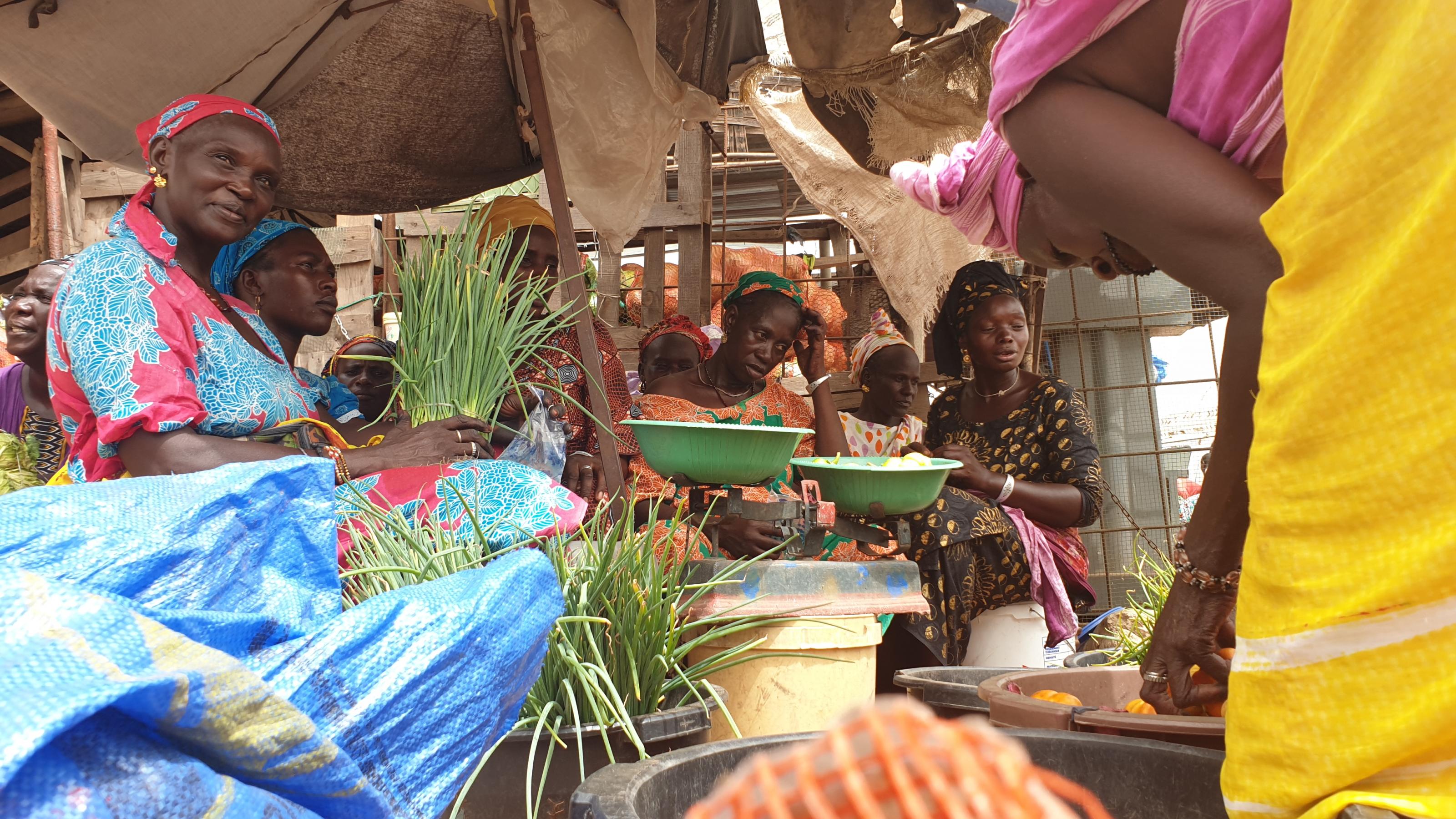 mehrere afrikanische Frauen in bunter Kleidung sitzen mit auf einem Markt unter einem Sonnenschirm, vor sich verschiedene Gemüse.