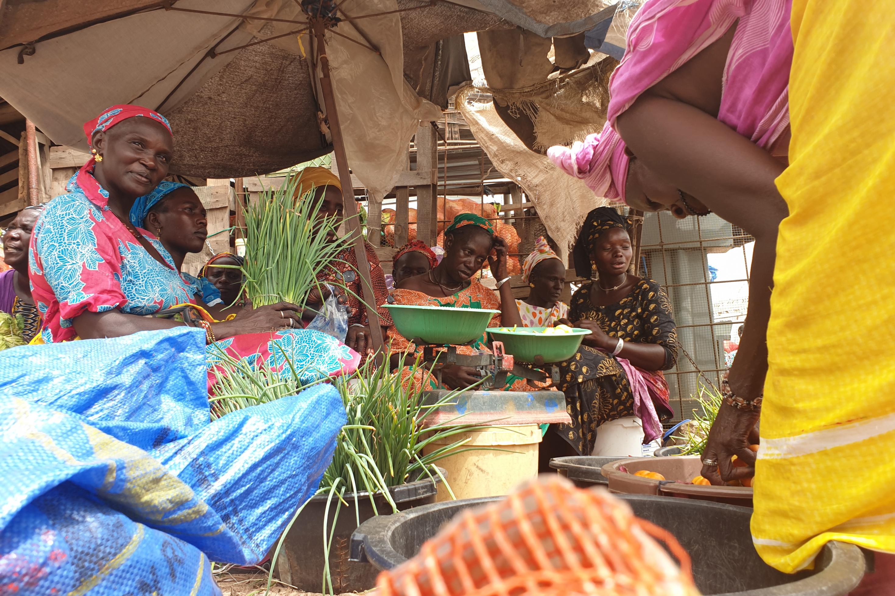 mehrere afrikanische Frauen in bunter Kleidung sitzen mit auf einem Markt unter einem Sonnenschirm, vor sich verschiedene Gemüse.