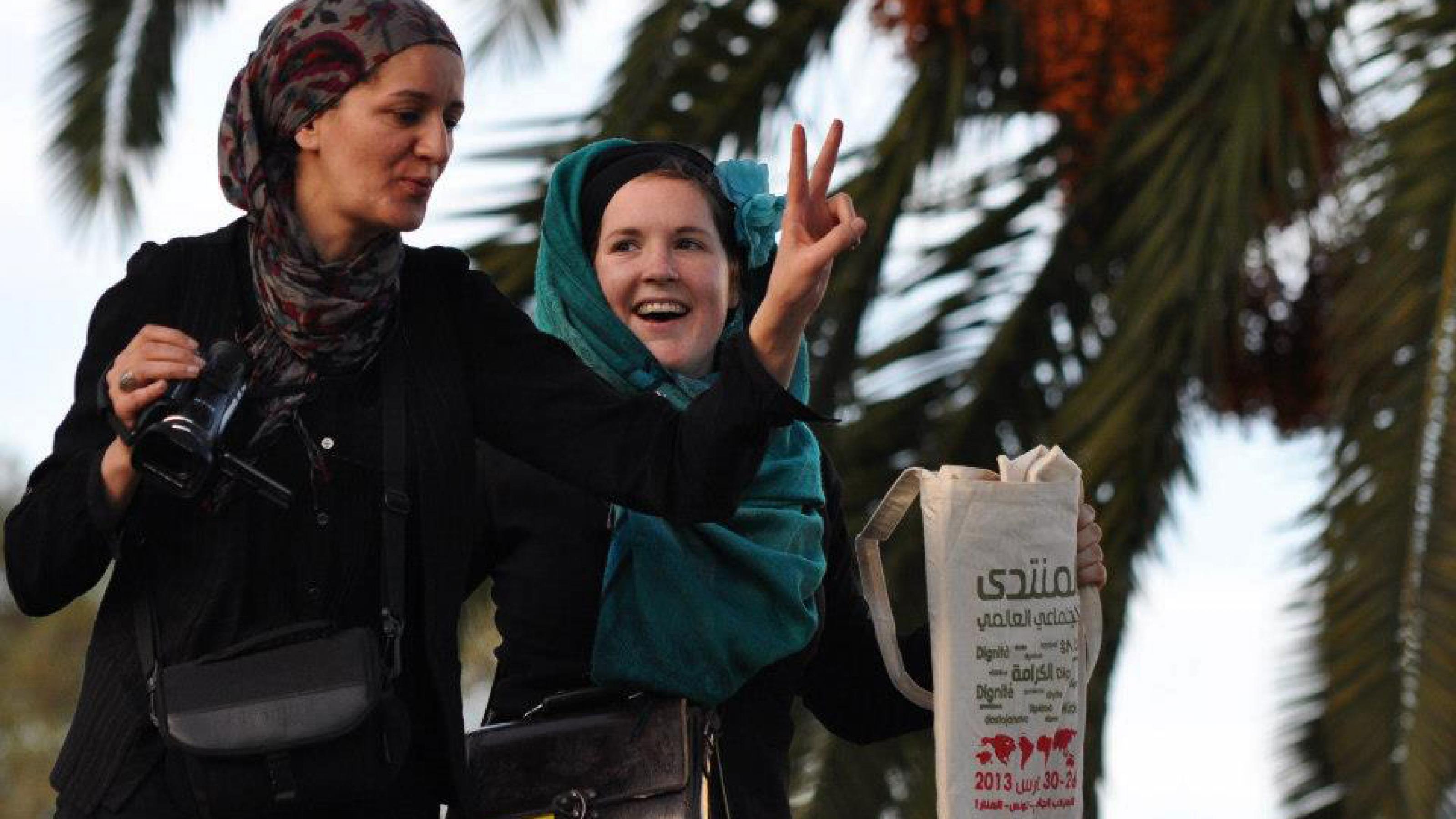Zwei Frauen, die in Tunis, Tunesien, auf einer Demonstration auf das Dach einer Bushaltestelle geklettert sind. Eine Frau hält eine Kamera in der Hand und macht mit der anderen das Victory-Zeichen, die andere halt einen Stoffbeutel mit dem Logo des Weltsozialforums hoch.