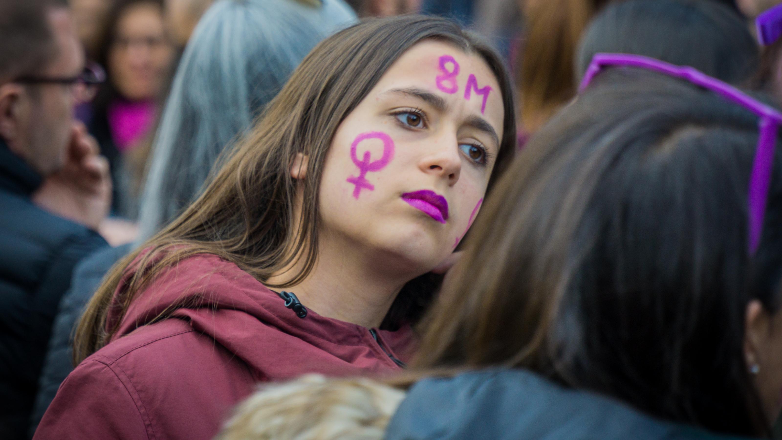 Inmitten einer Menschenmenge blickt eine junge Frau mit Frauenzeichen und 8M auf der Wange und lila geschminkten Lippen entschlossen in die Ferne.