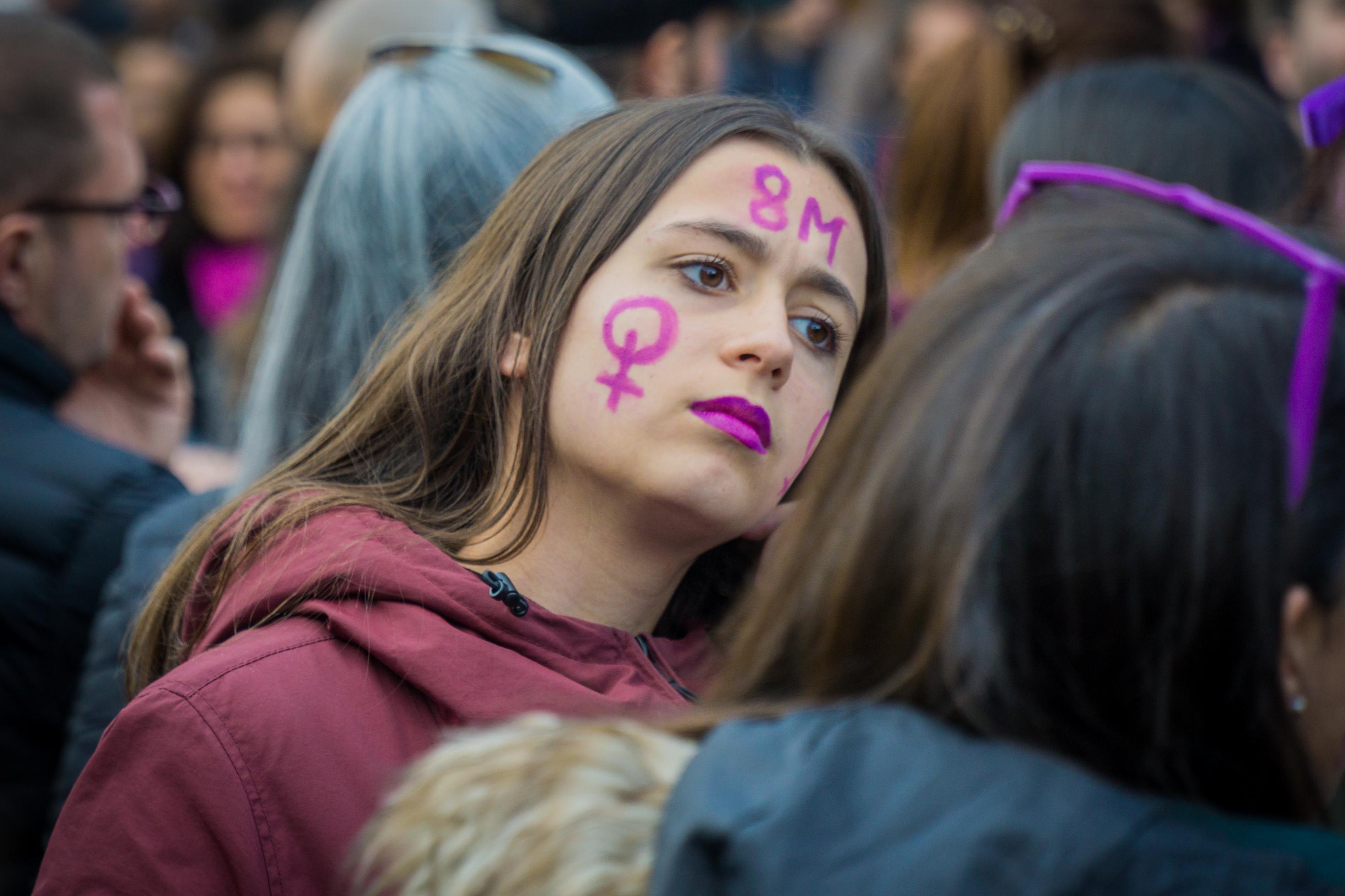 Inmitten einer Menschenmenge blickt eine junge Frau mit Frauenzeichen und 8M auf der Wange und lila geschminkten Lippen entschlossen in die Ferne.